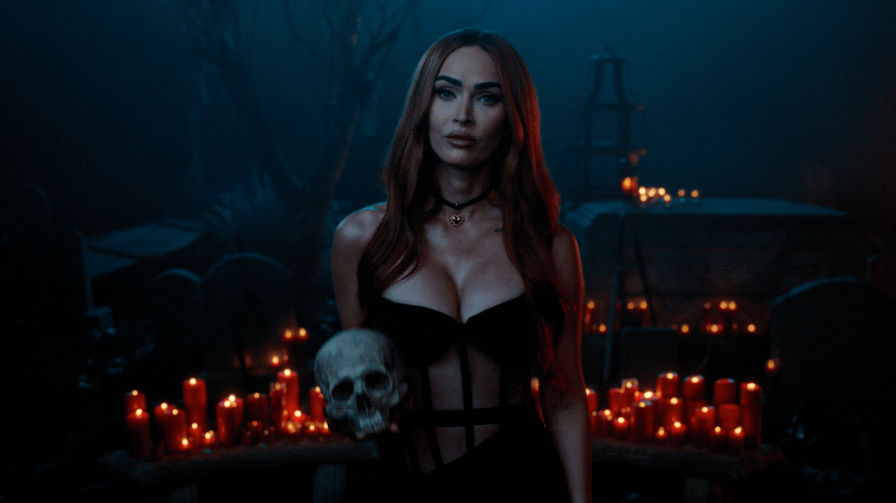 A woman with long red hair and dark makeup holding a skull in a dimly lit setting with red candles, skulls, and dark decorations.