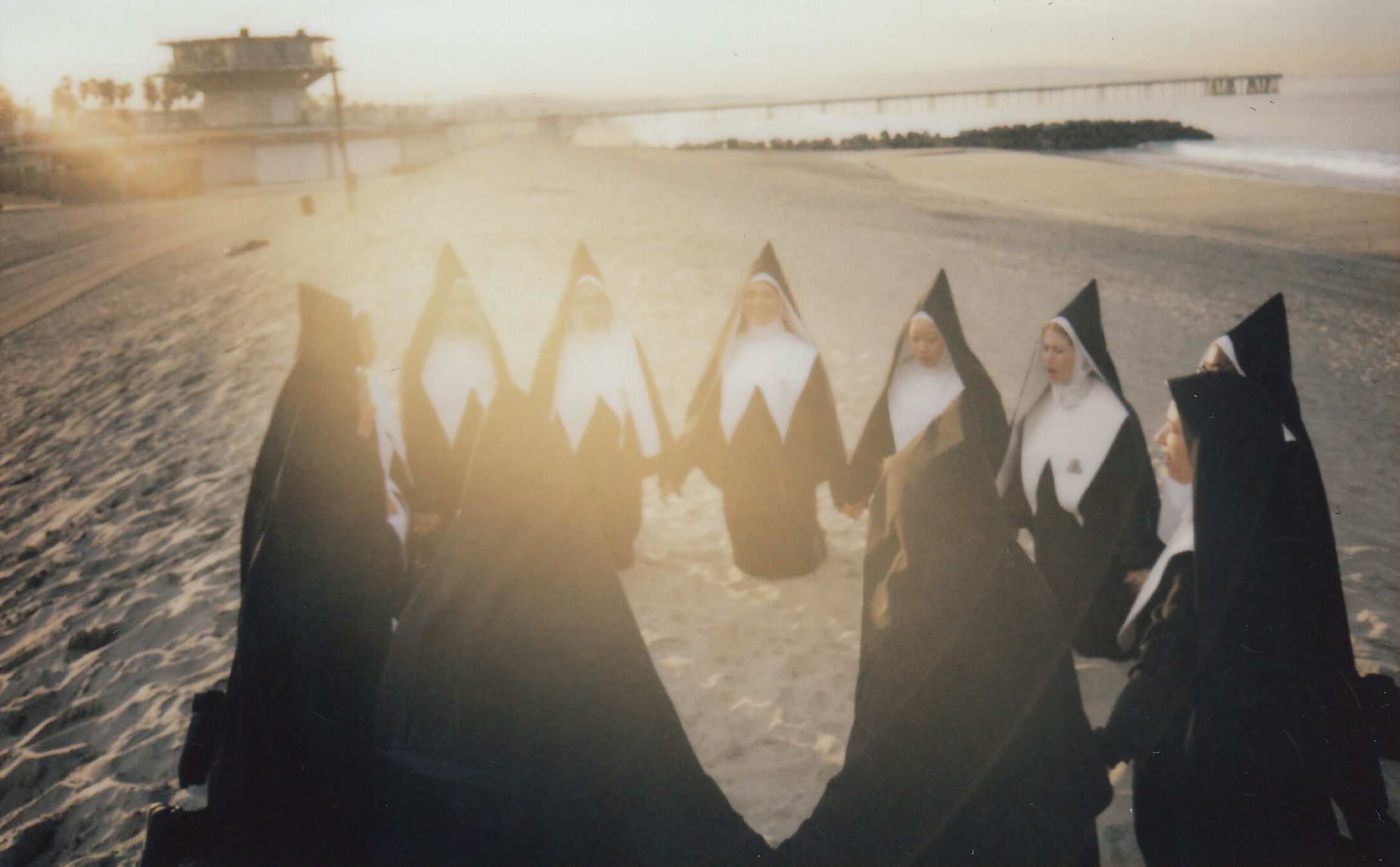 A group of women dressed as nuns, holding hands in a circle on a beach at sunset.