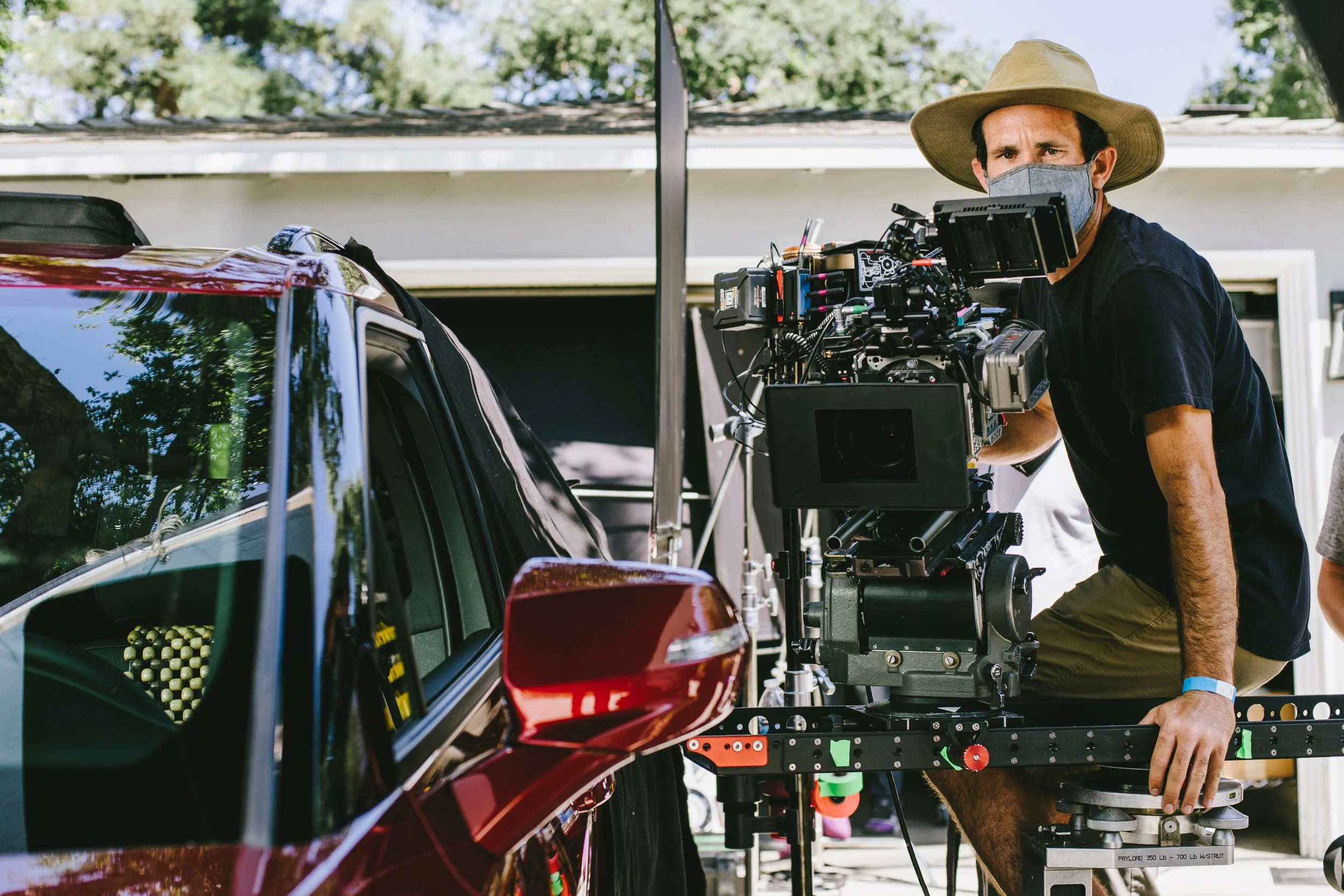 Filmmaker operating a professional camera mounted on a track, filming a red car outdoors during daytime.