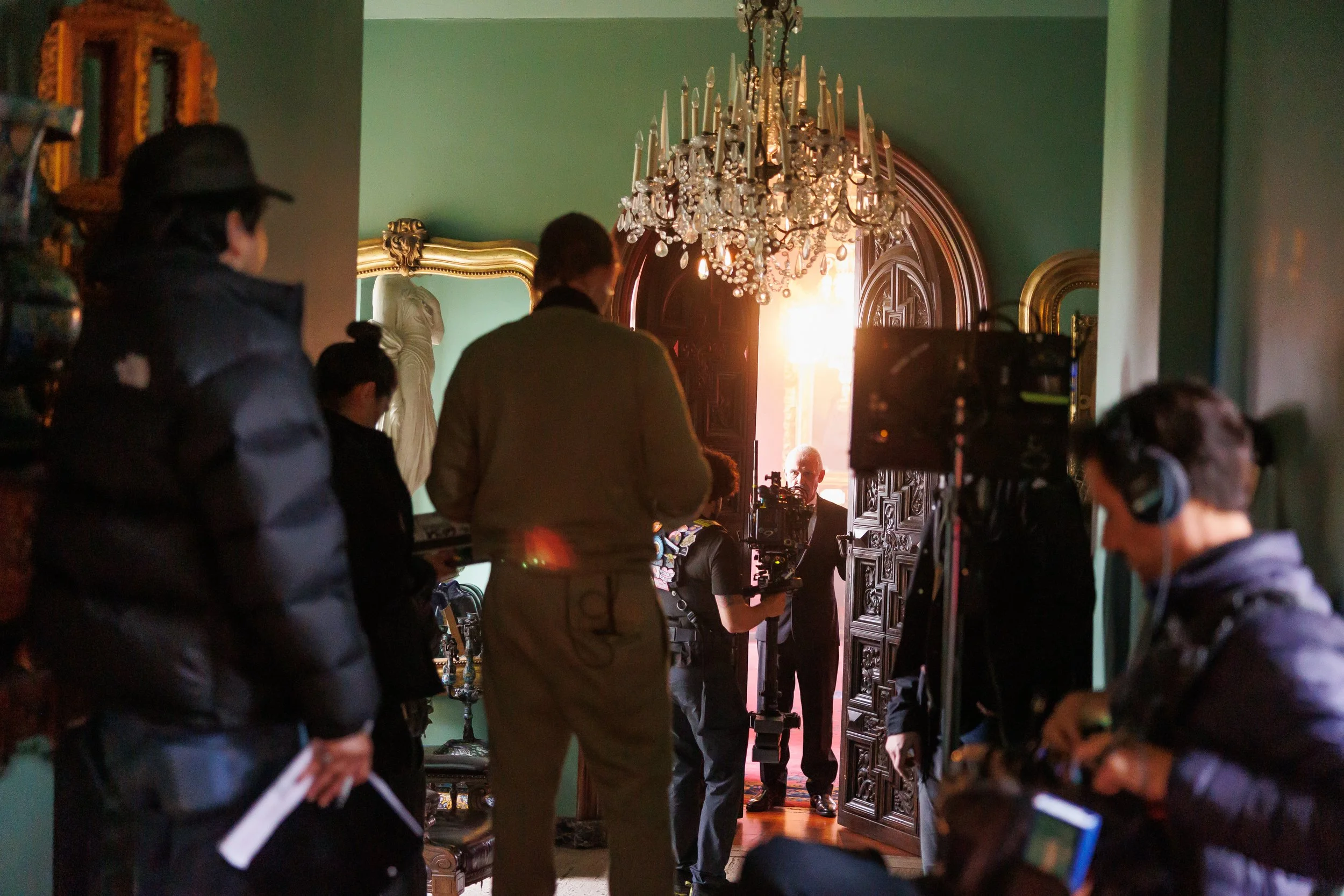 A film crew is filming scenes inside a historic or elegant house, with a man in a suit on camera in front of a large ornately carved wooden door, illuminated by natural light from the doorway. Crew members with equipment and headphones surround him, 