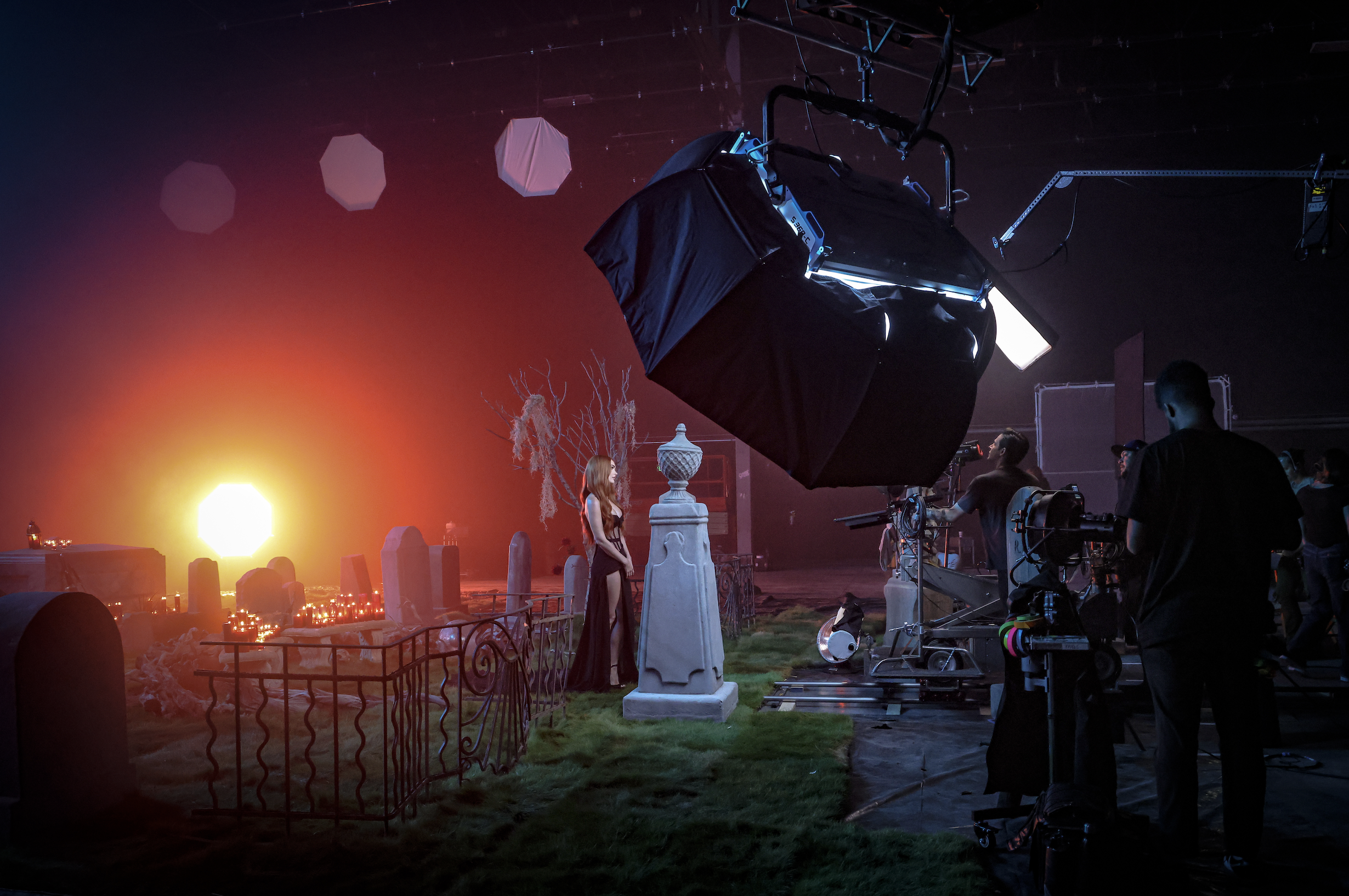 Behind the scenes of a film or music video shoot with a set resembling a graveyard, featuring a woman in a black dress, candles, and tombstones, with professional lighting and camera equipment, and a fiery sunset or explosion in the background.