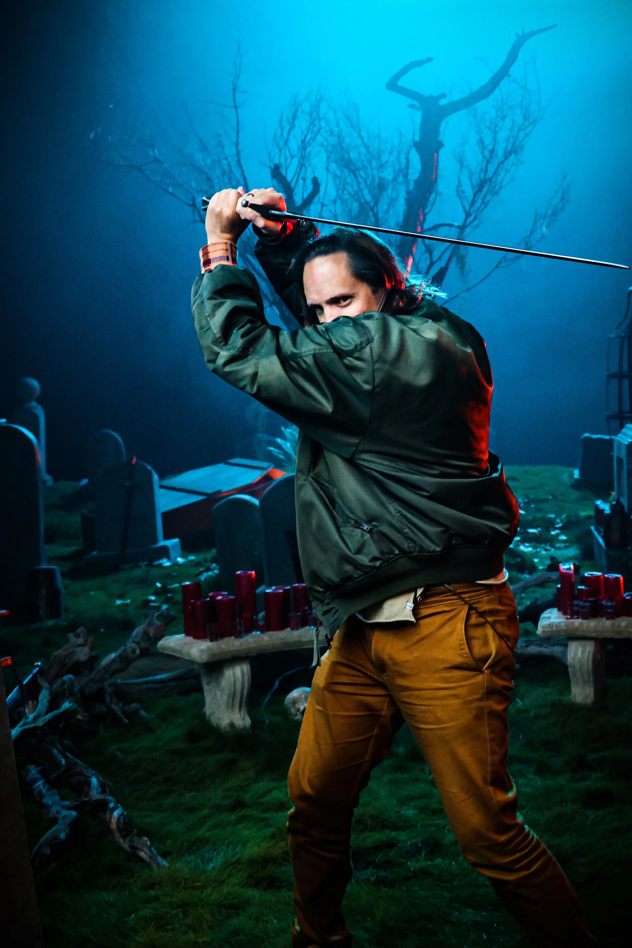 A person with long dark hair, wearing a green jacket and brown pants, striking a fighting pose with a sword in a spooky cemetery scene with tombstones, candles, and a leafless tree in the background, illuminated by blue light.