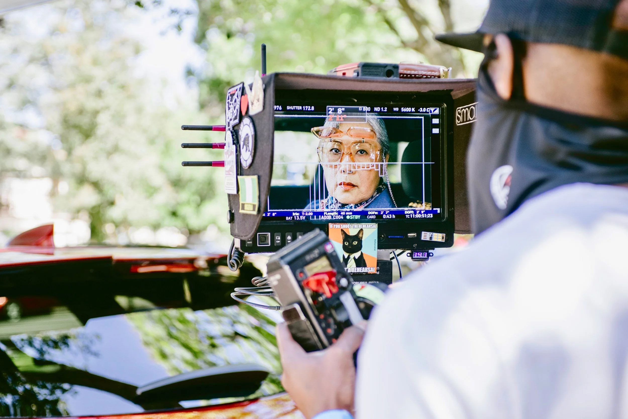 A film director operating a camera on a sunny outdoor set features an older woman actor with gray hair, glasses, and a patterned scarf, looking into the camera.
