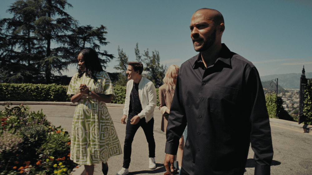 Group of four people walking outdoors on a sunny day, with greenery and cityscape in the background, dressed casually.