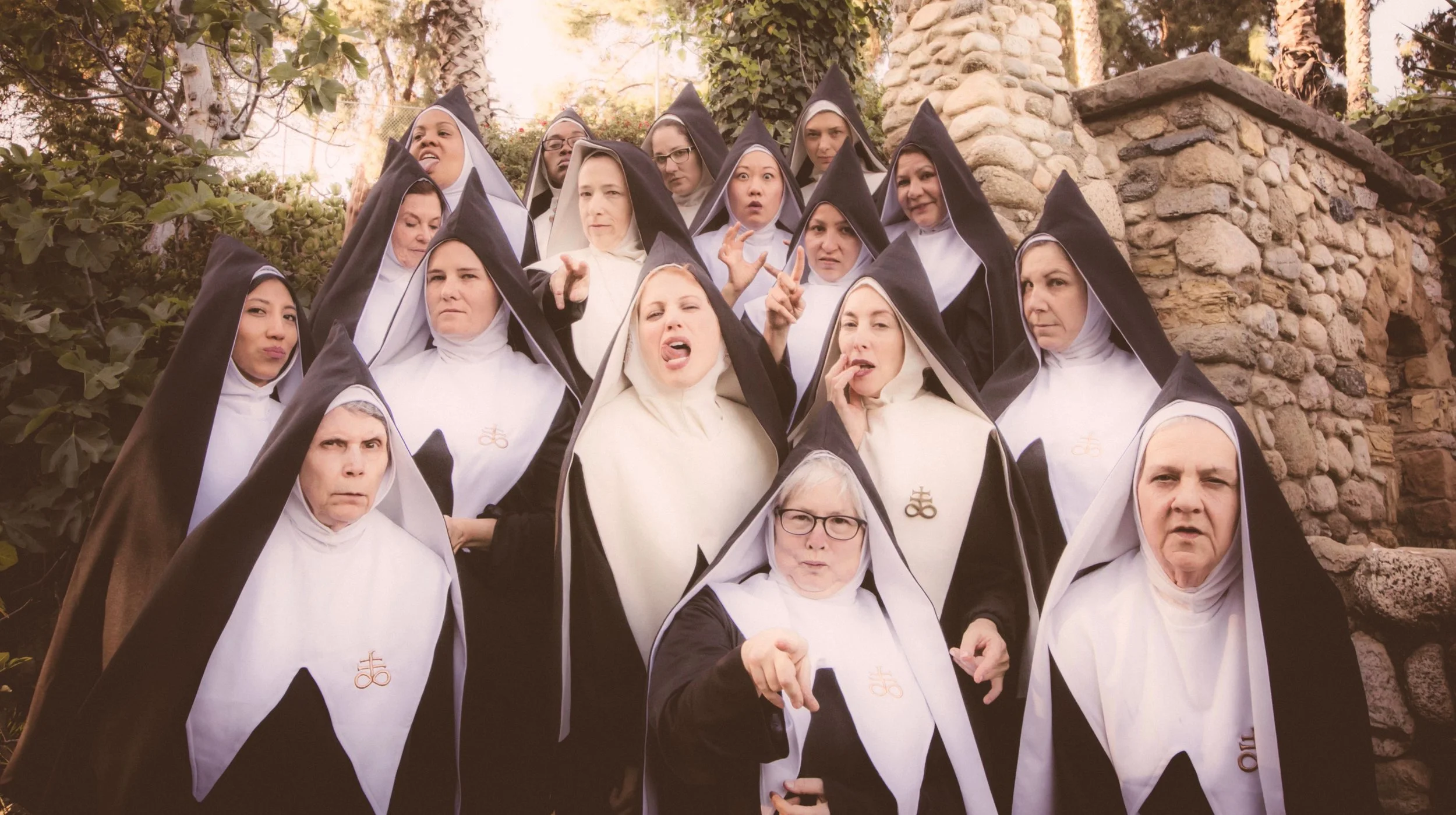 Group of women dressed as nuns in black and white habits making various facial expressions and gestures outdoors near a stone wall and trees.