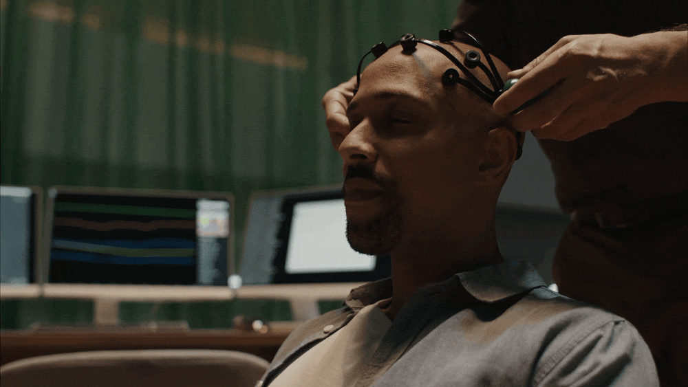 A man is sitting in a room with multiple monitors, wearing a cap with sensors, while another person attaches or adjusts the sensors on his head. The man appears to be getting a brain scan or EEG test.