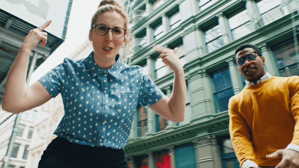 Two people in an urban setting with tall buildings, one woman in a blue polka dot shirt and glasses pointing and speaking, and a man in a mustard yellow jacket behind her.