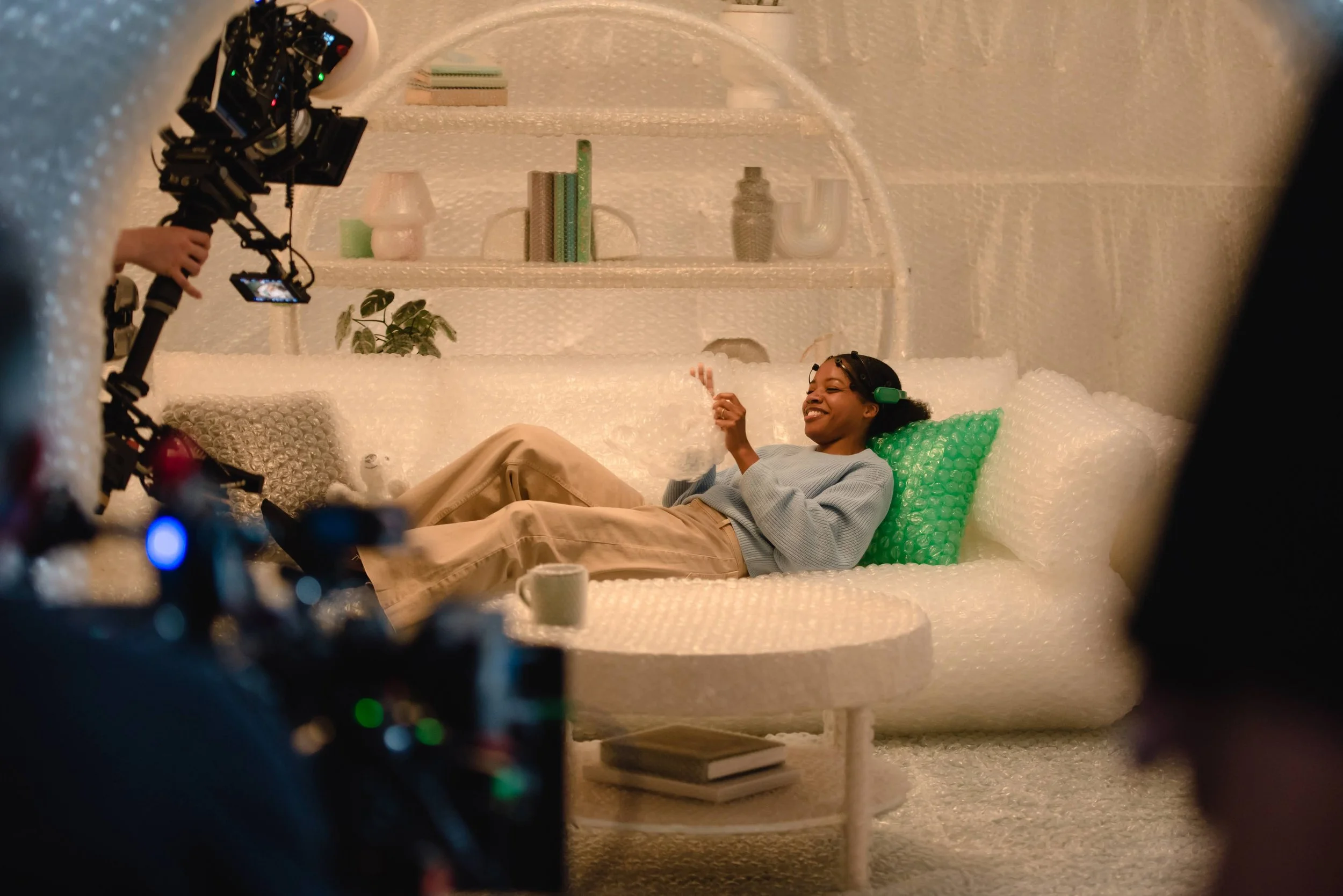 A woman lying on a white textured couch with a green pillow, smiling, with large headphones on, in a cozy, modern room with shelves, books, plants, and decorative objects, while being filmed by a professional camera.