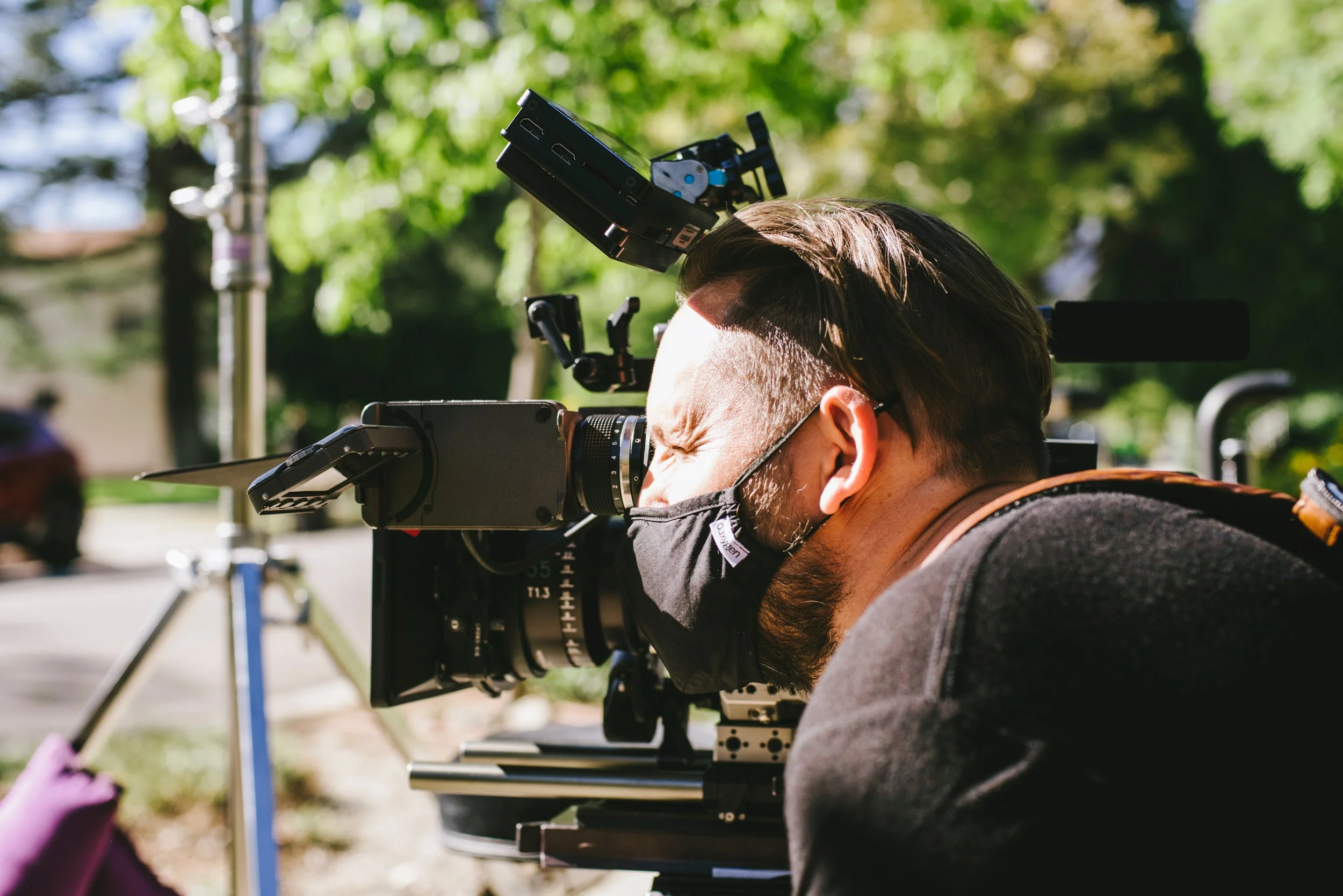 Person aiming a professional film camera outdoors, wearing a black face mask and with sunlight on their face.