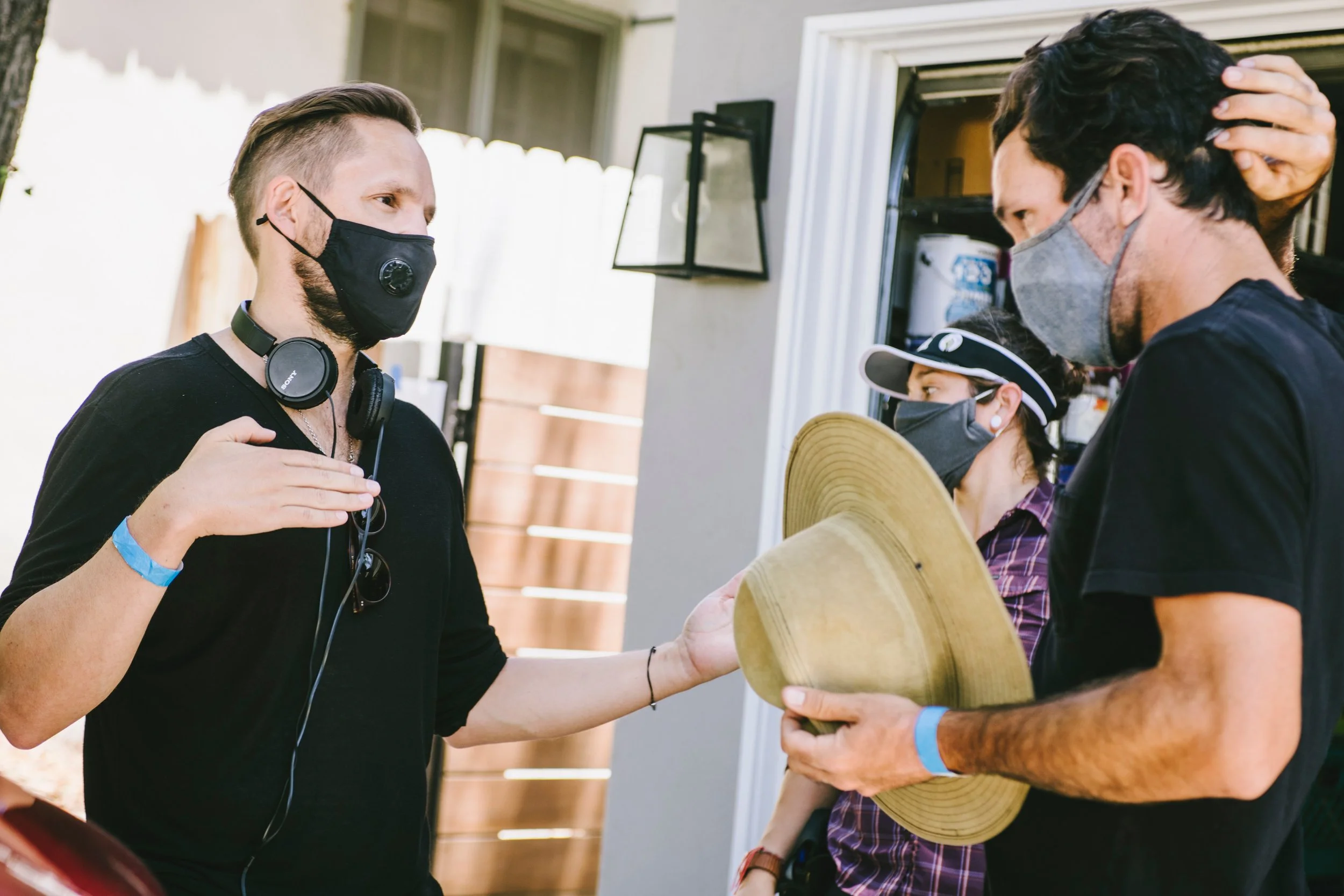 Three people engaged in a conversation outdoors, all wearing protective masks. One person has headphones around his neck and is touching his chest, another is holding a large straw hat, and the third person, a woman, is wearing a visor and sunglasses