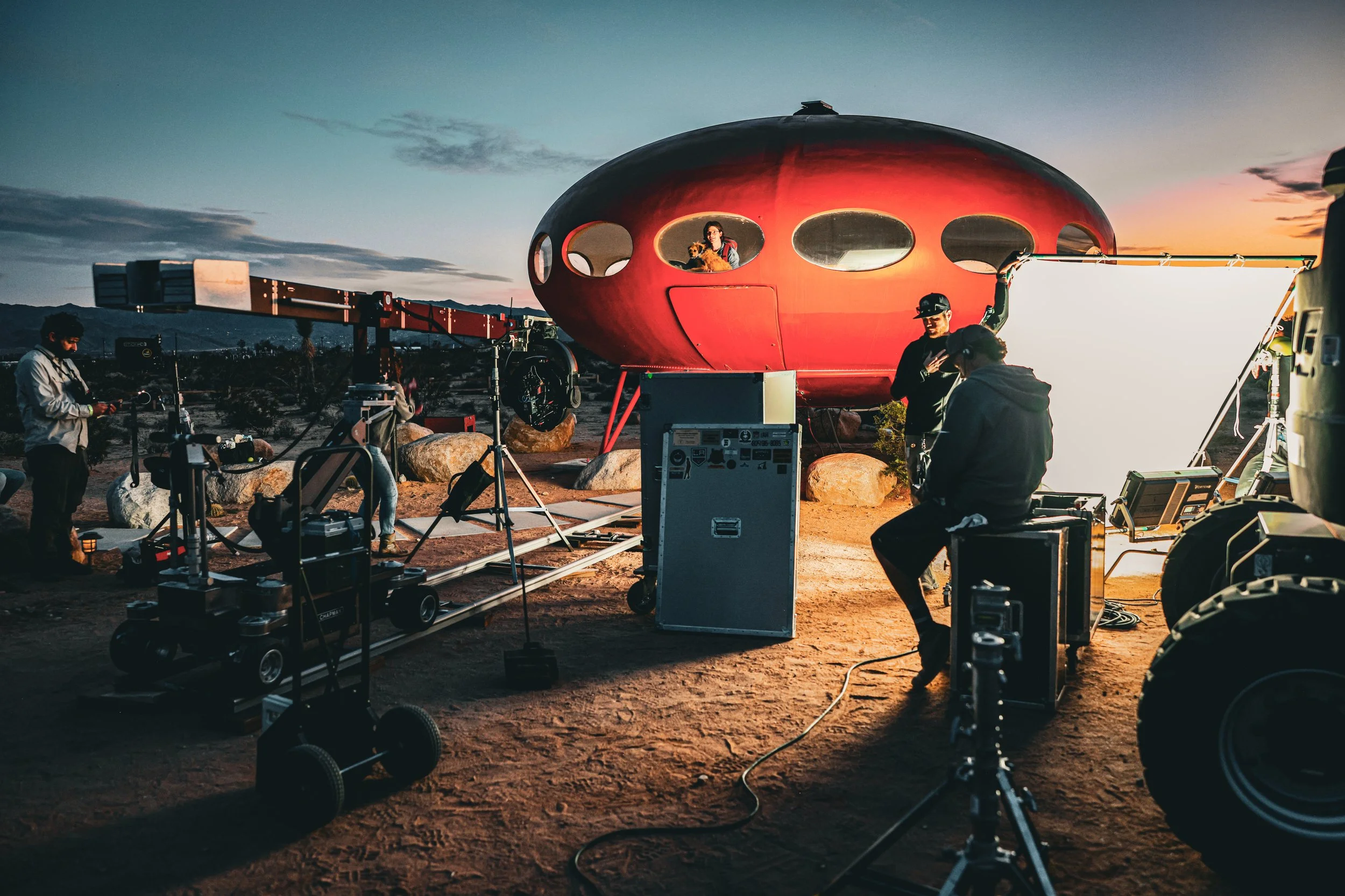 Film crew setting up a shoot around a red, futuristic spaceship-like structure during sunset in a desert landscape.