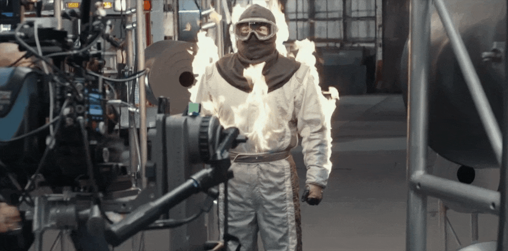 Person standing in front of a film camera, dressed in white protective suit, goggles, and face mask, with flames in the background.