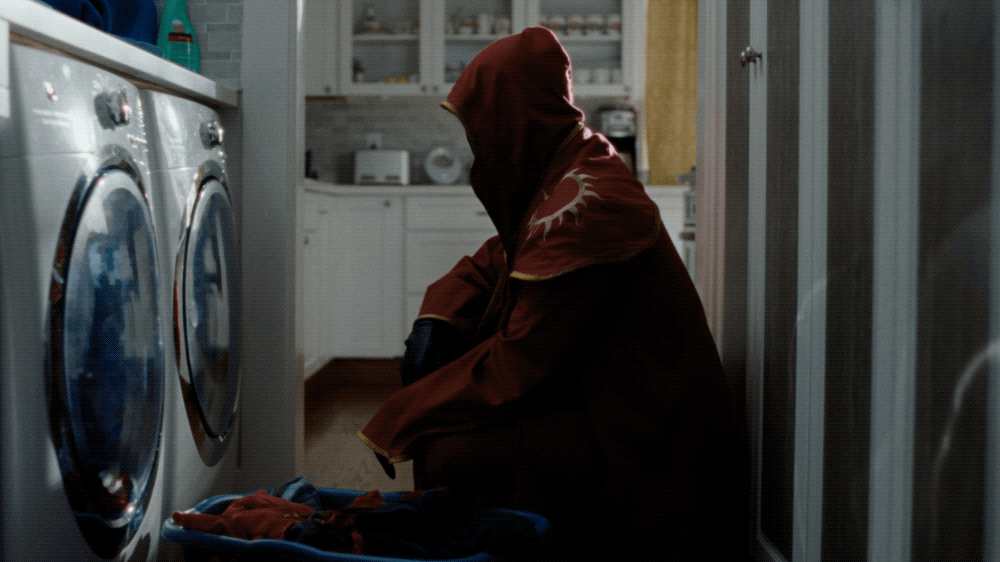 Person wearing a hooded jacket with a sun design on the hood, sitting on the floor of a laundry room, next to a washing machine.
