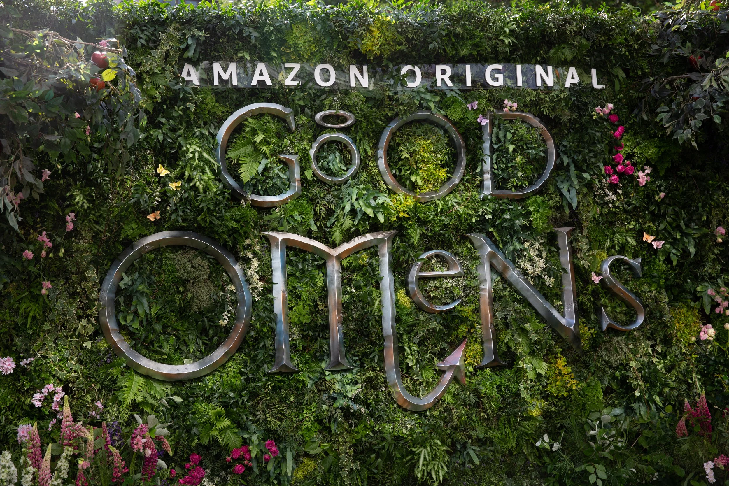 Green wall decorated with various plants and flowers with the words 'Amazon Original Good Omens' in metal lettering.