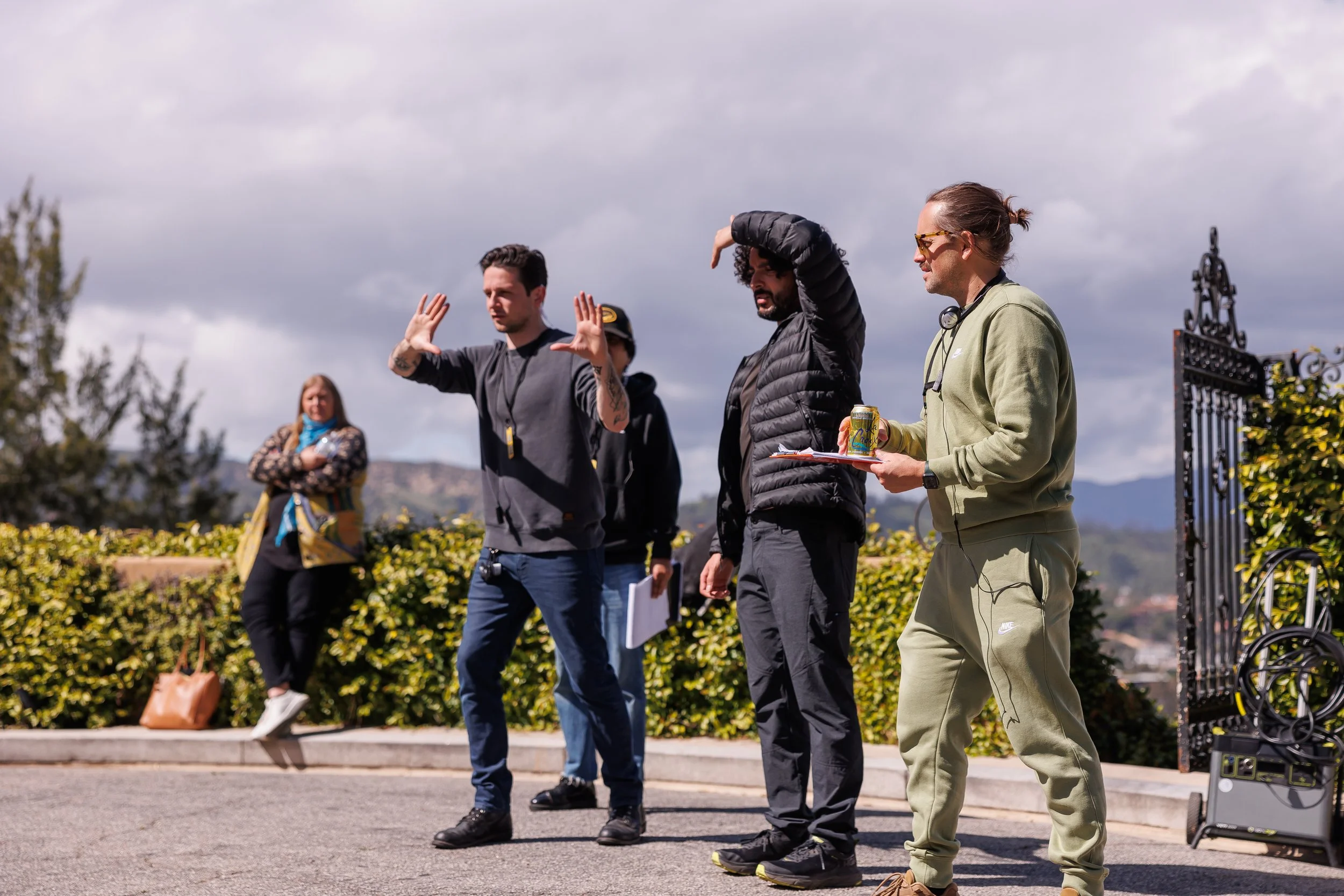 Group of five people standing outdoors, three men in the front and two women in the background, with cloudy sky and greenery in the distance. The man on the left is gesturing with his hands, the man in the center is shading his eyes from the sun, and