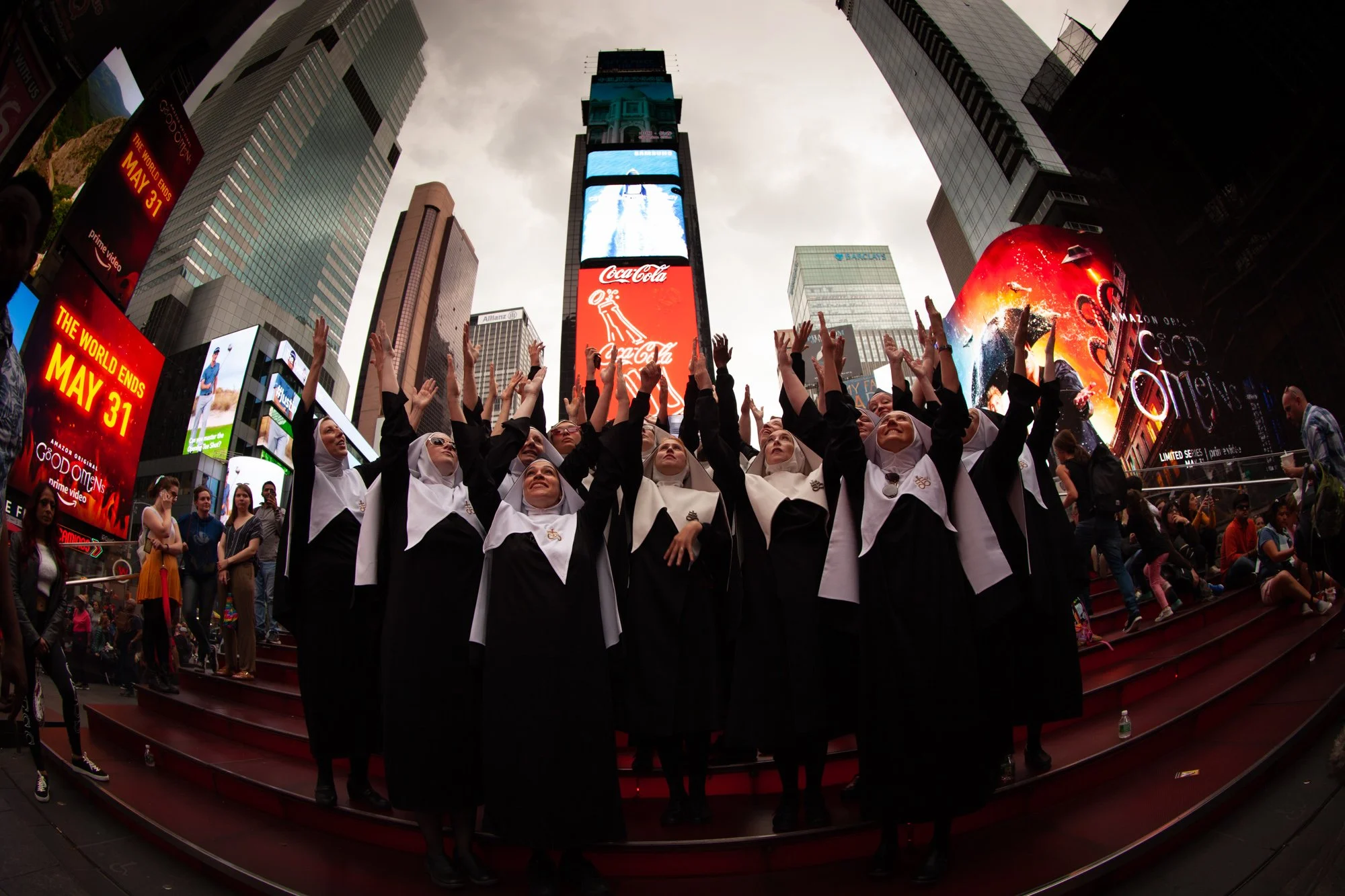 Group of women in black and white graduation gowns and caps, raising their hands in celebration, on red steps in Times Square, New York City, with digital billboards and skyscrapers in the background.