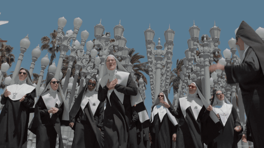A group of nuns in black and white habits walking outside, with tall decorative lamp posts and palm trees in the background under a blue sky.