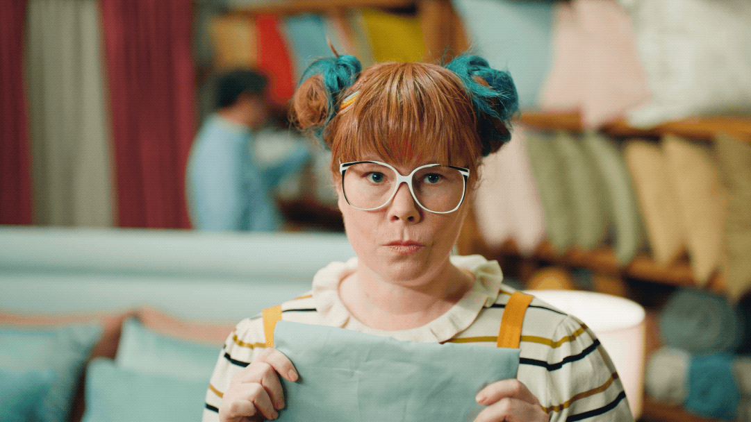 A young woman with red hair, glasses, and rainbow-colored hair buns is holding a fabric sample in a fabric store.