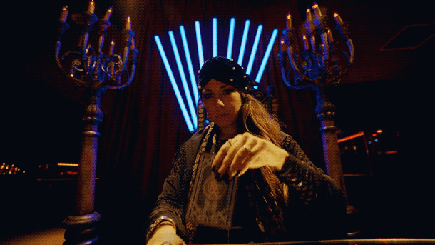 Woman wearing a hat and gloves sitting at a table in a dimly lit room with neon light decorations and chandeliers.