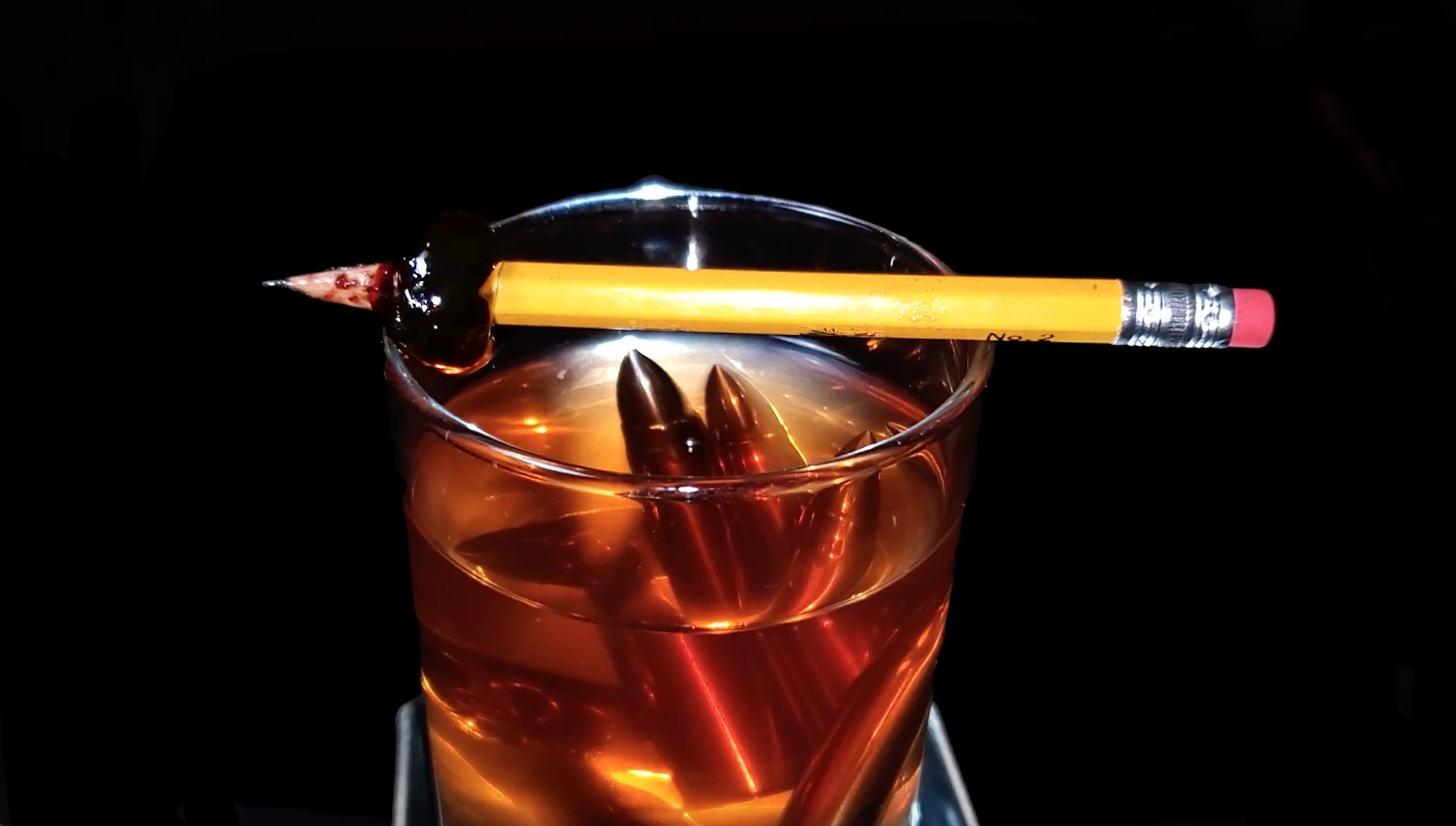 A glass of amber-colored beverage with ice, topped with three bullets, a cherry, and a yellow pencil with a red eraser, against a dark background.