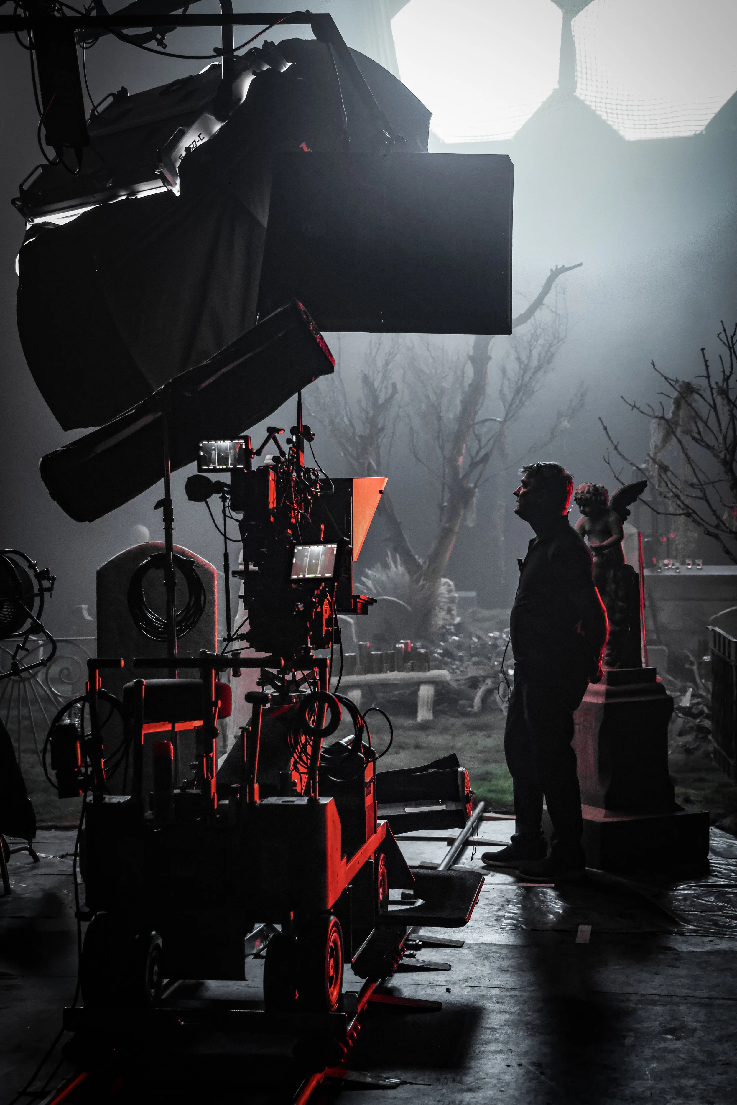 Filmmaker in silhouette on set with professional camera equipment, behind scenes of a spooky outdoor scene with leafless trees and a sculpture of a winged figure.