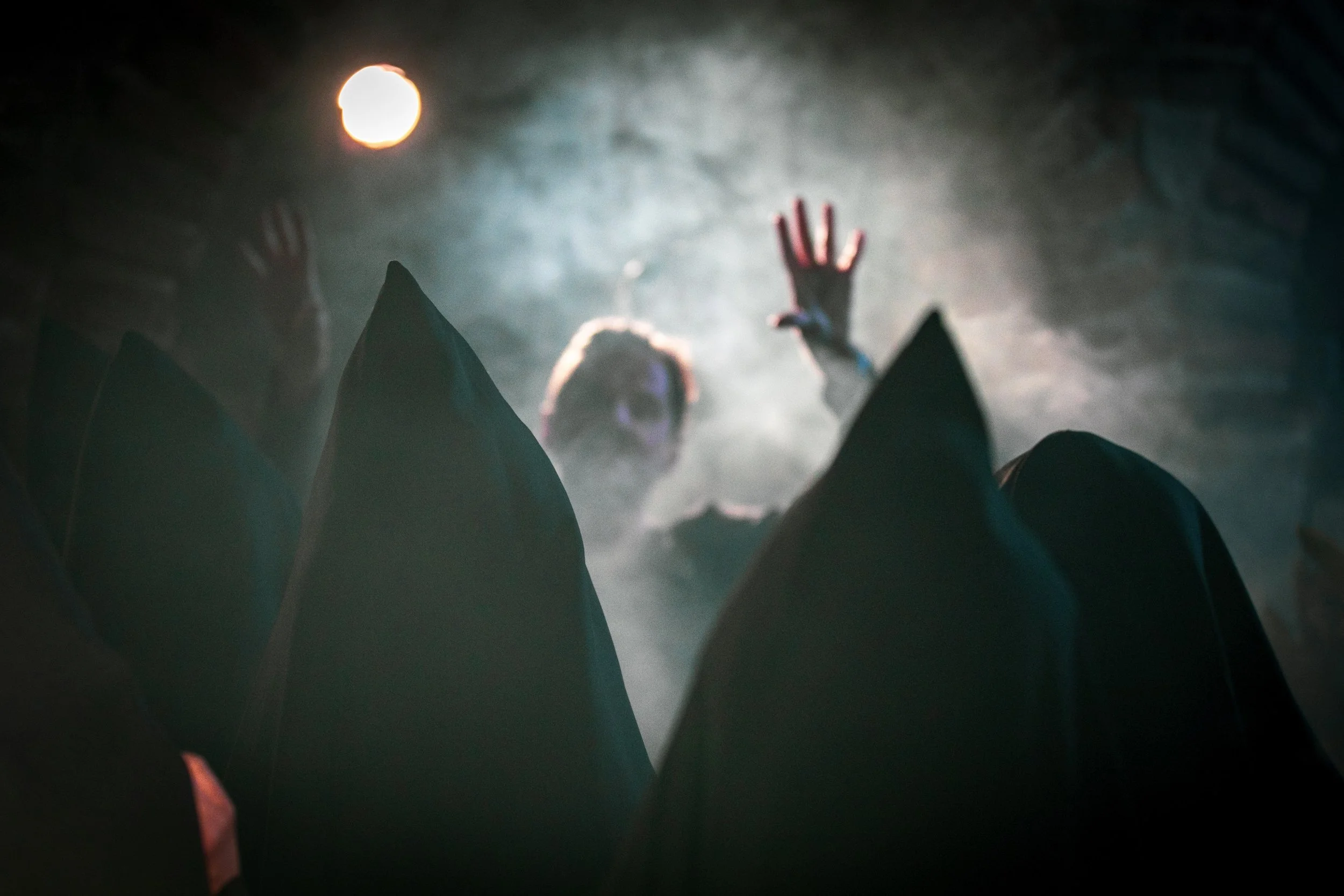 A person dressed as a witch or wizard with a pointed hat is reaching out with their hand towards the camera, surrounded by fog and smoke, with a full moon in the background. Good Omens Amazon Hal Kirkland