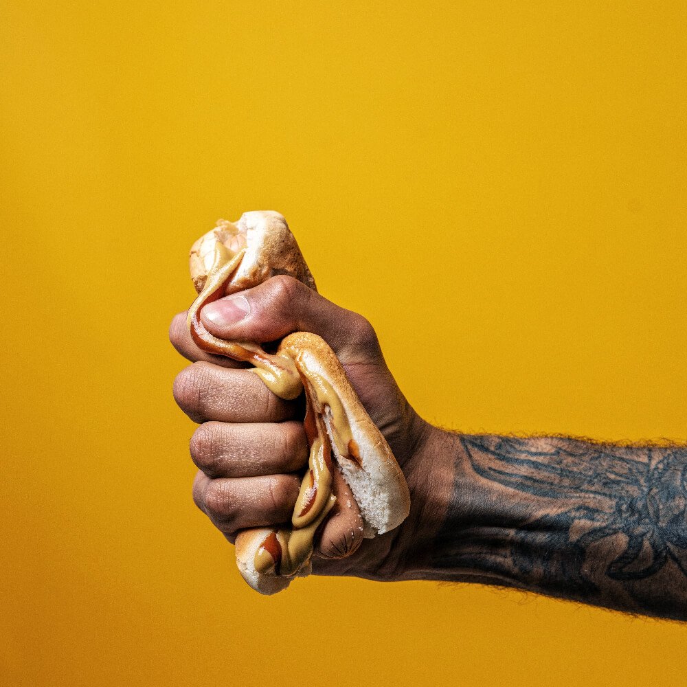 a tattooed arm reaches out from the side panel of this hyper-stylized food photo, enthusiastically squeezing a ketchup and mustard-dressed hotdog until it's just become an absolute mess. I don't know why the person does this, but it's really funny