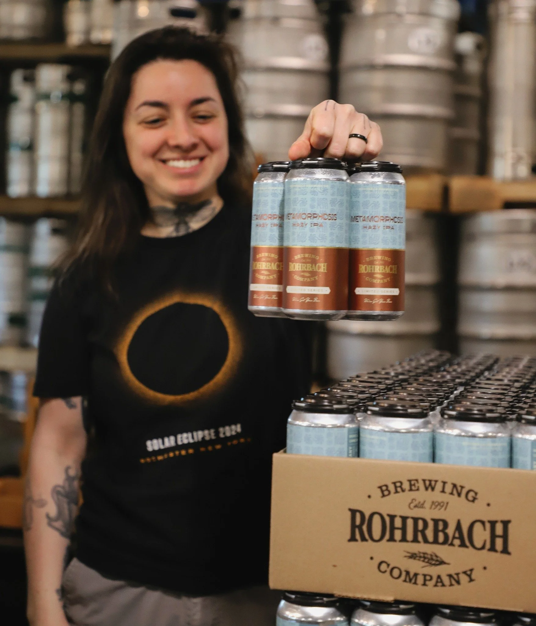 brewer in the brewery holding a 4-pack of the latest limited release, Metamorphosis IPA, with a blue and brown label