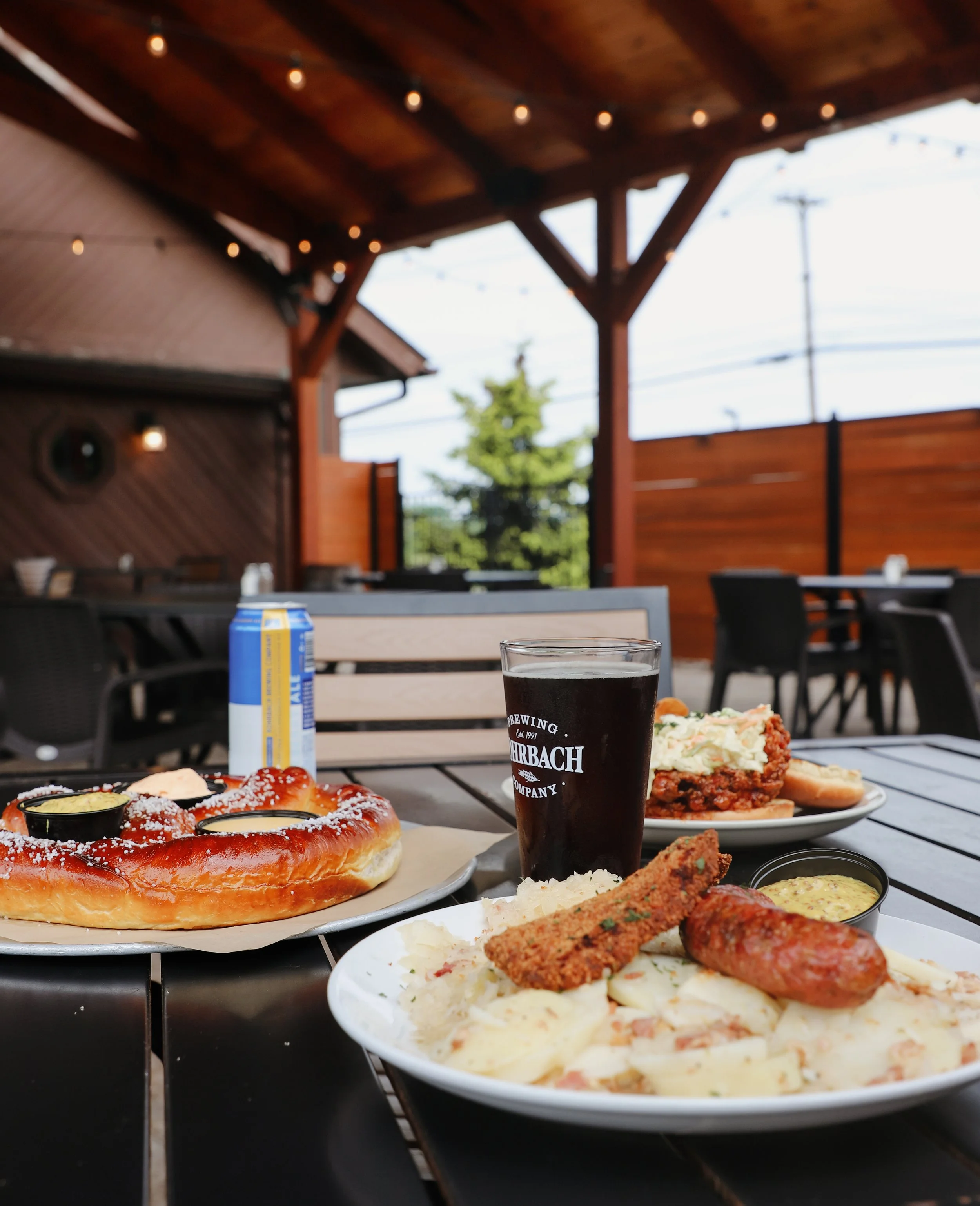 Outdoor dining area with wooden roof, string lights, and a view of trees and power lines in the background, featuring plates of pizza, potato salad, fried chicken, and drinks on the tables.