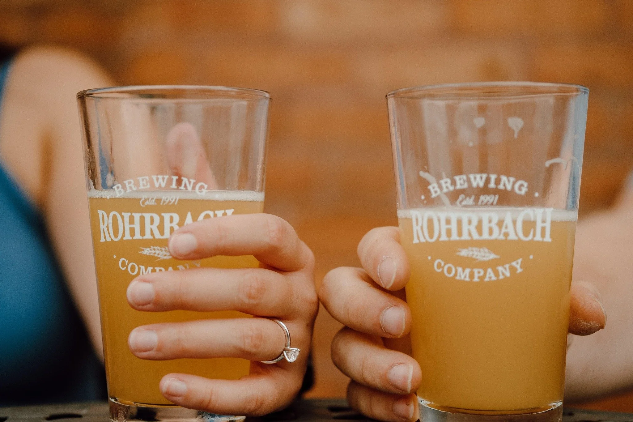 Two hands holding clear beer glasses with the logo 'Rohrbach Brewing Company' printed on them, filled with beer, against a blurred warm background.