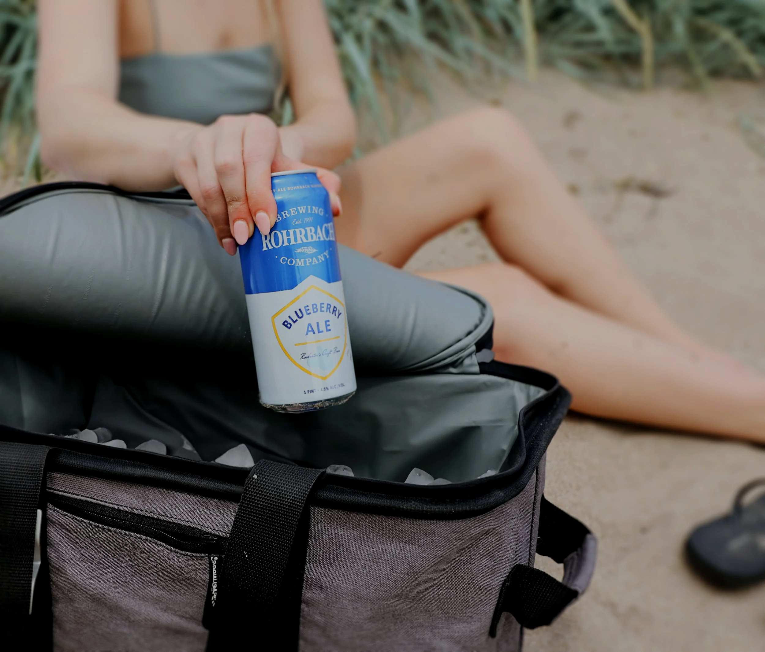 Person holding a can of Rohrbach Blueberry Ale over an ice-filled cooler at the beach.