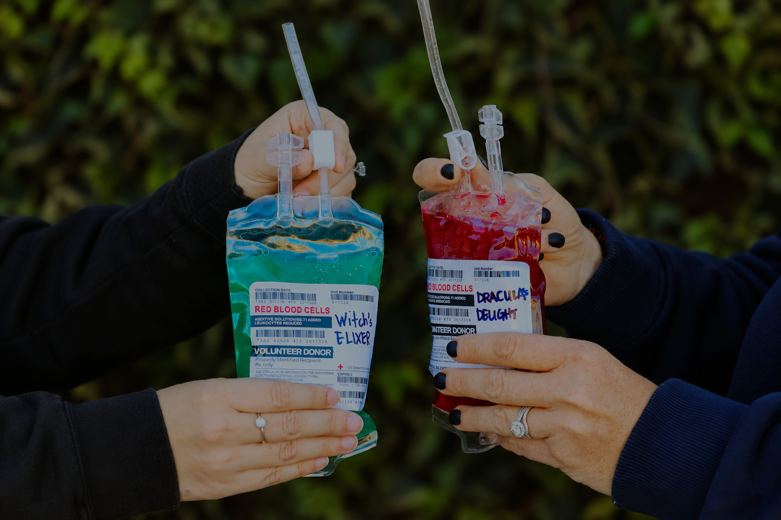 Two people hold colorful Halloween drinks in IV bags labeled Witch’s Elixir and Dracula’s Delight.