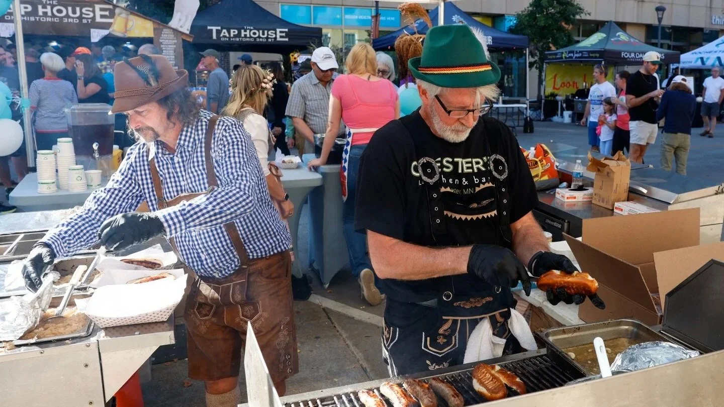 Two men in traditional German attire grill sausages and pretzels at an outdoor festival.