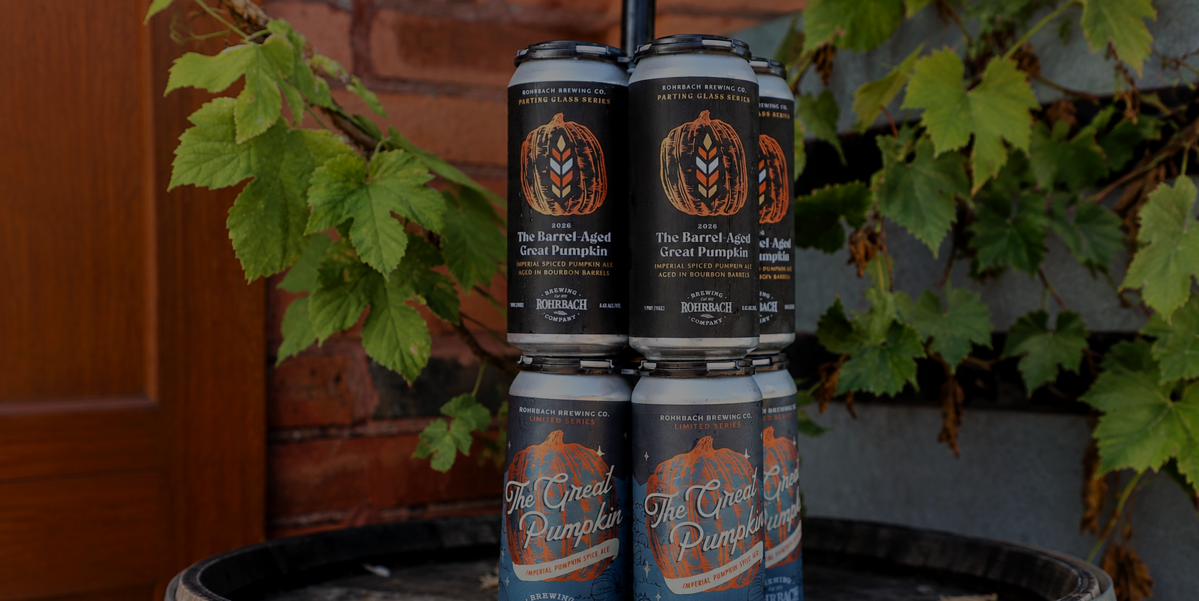 Rohrbach Great Pumpkin and Barrel-Aged Great Pumpkin cans stacked on a barrel with fall greenery.