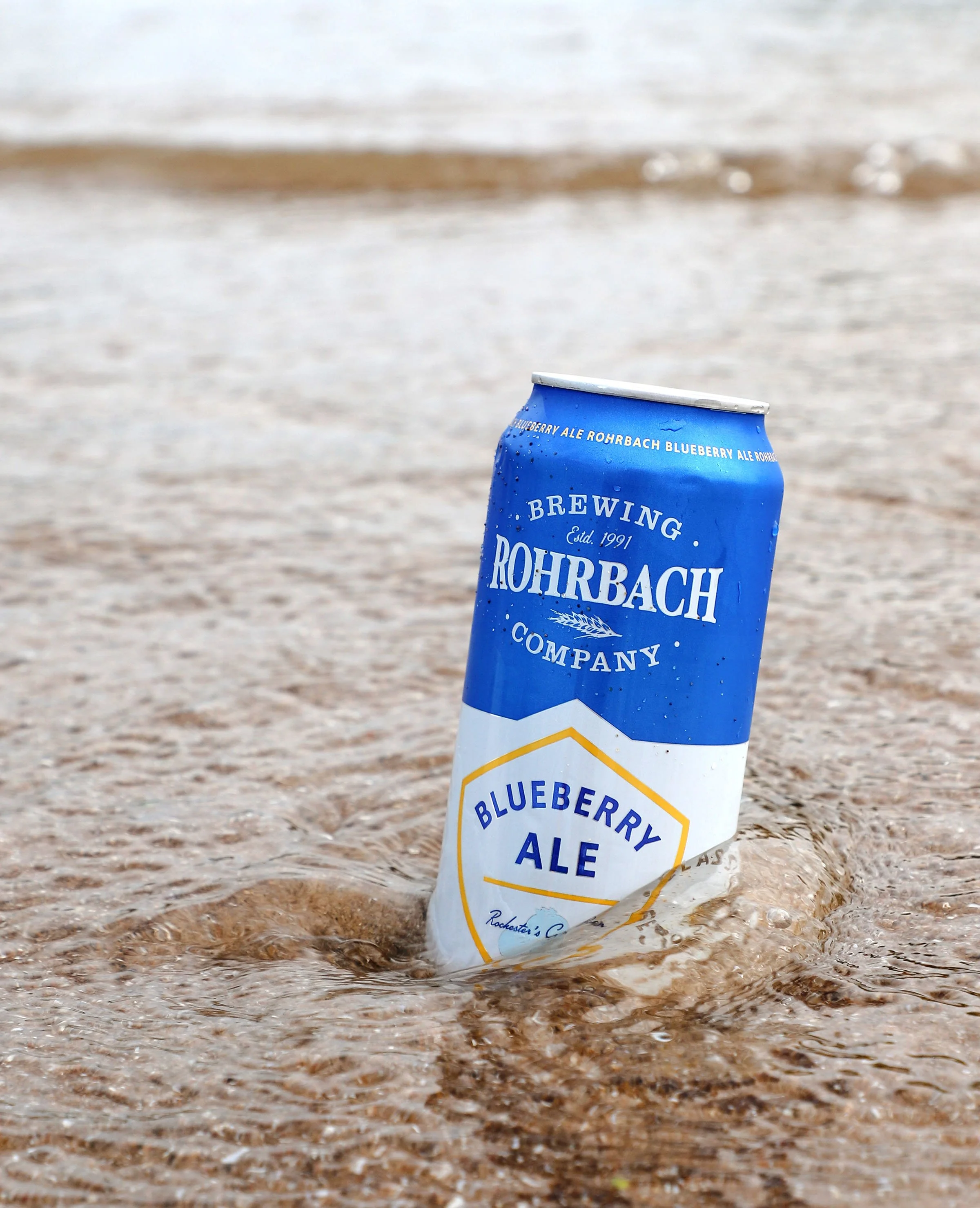A blue aluminum can with 'Blueberry Ale Rohrbach Brewing Company' written on it, partially submerged in water on a sandy beach.