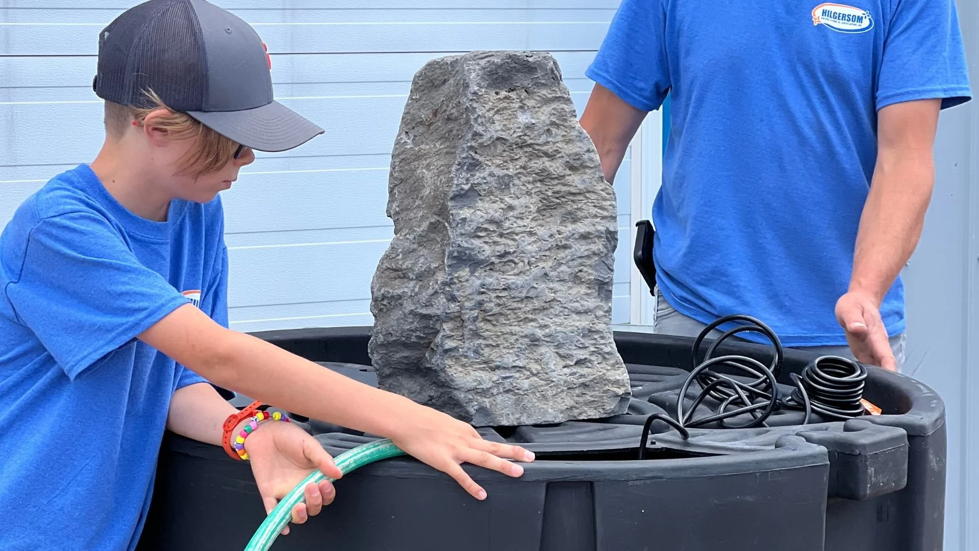 Jerry and his eldest child build a water feature using a grey boulder, black plastic tub, black tubing and a small pump