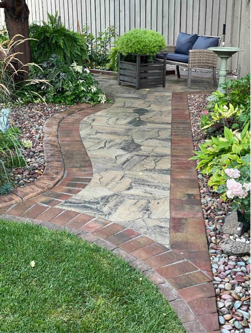 A cobbled pathway and patio with brick border is accented on either side with decorative gravel and lush perennials.