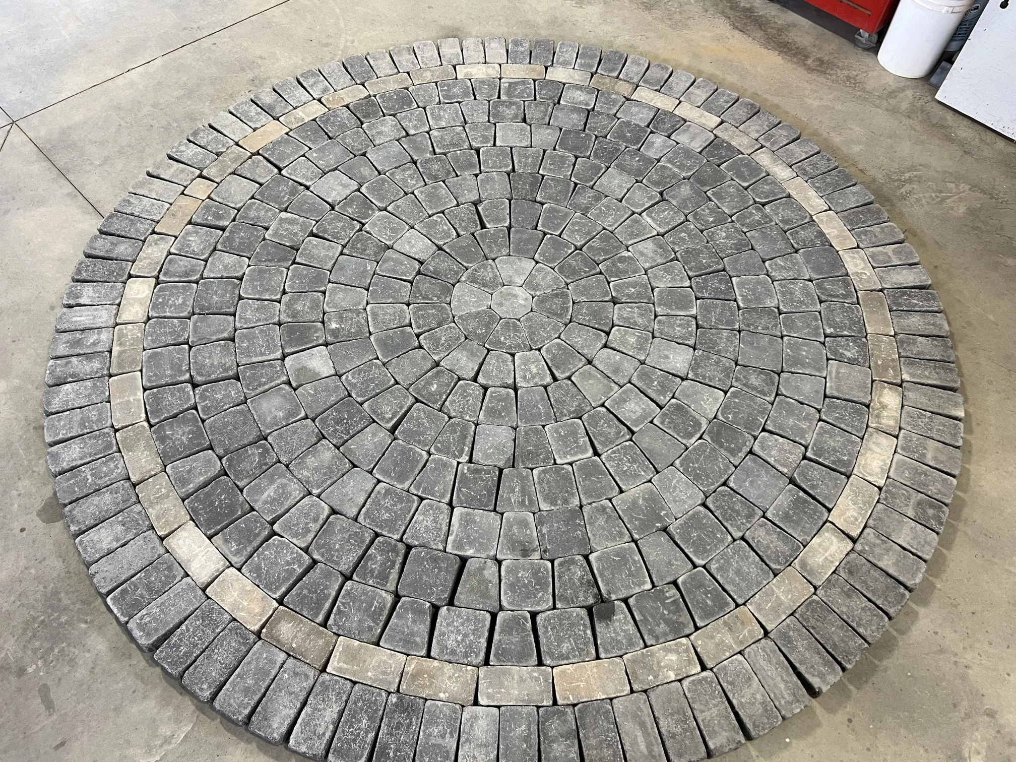 circle patio made from gray cobbled paving stone. As an accent, a single layer of taupe paving stones runs between the outer-most and inner-most part of the patio.