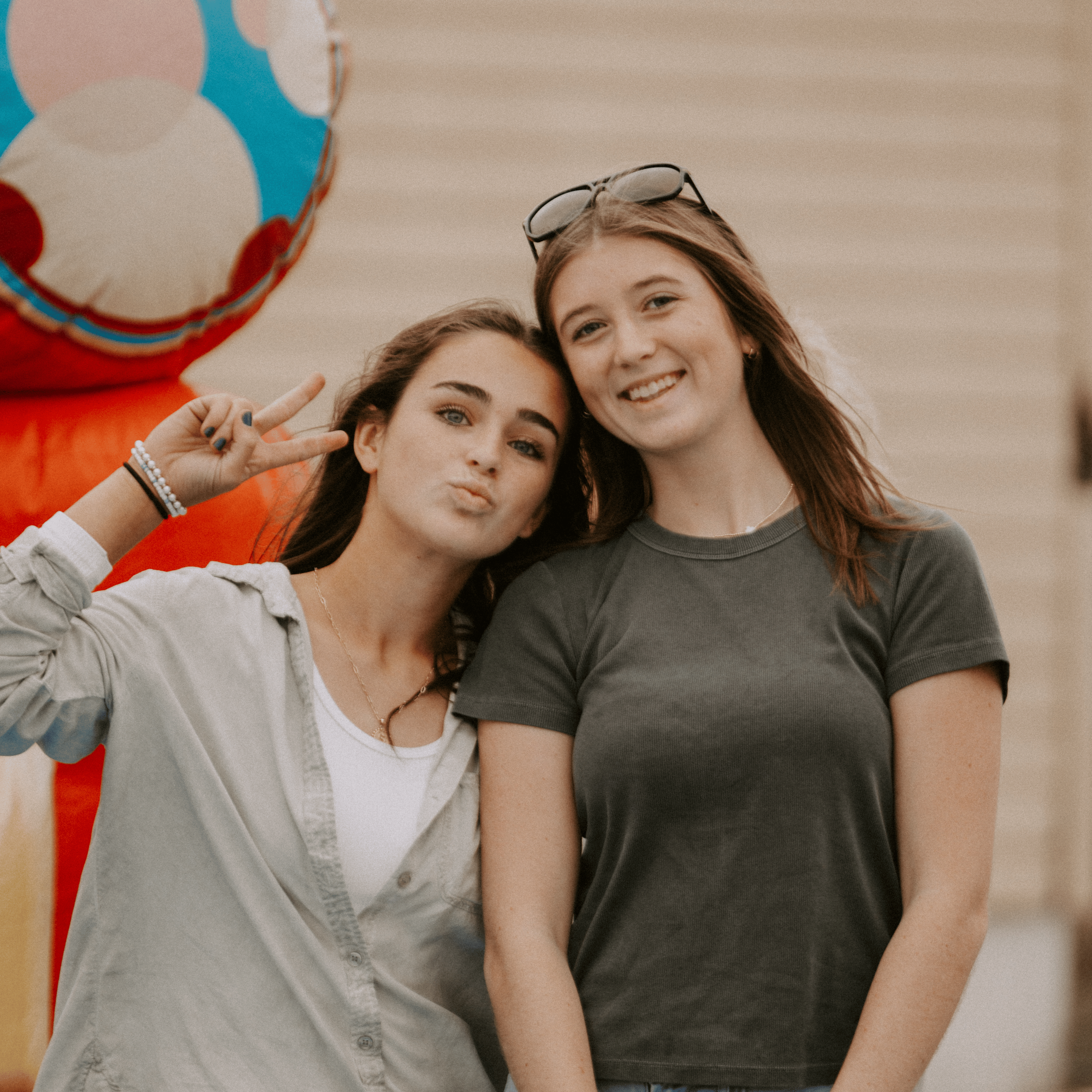 Two young women smiling and standing close together outdoors, with one woman wearing sunglasses on her head and the other making a peace sign near her face.