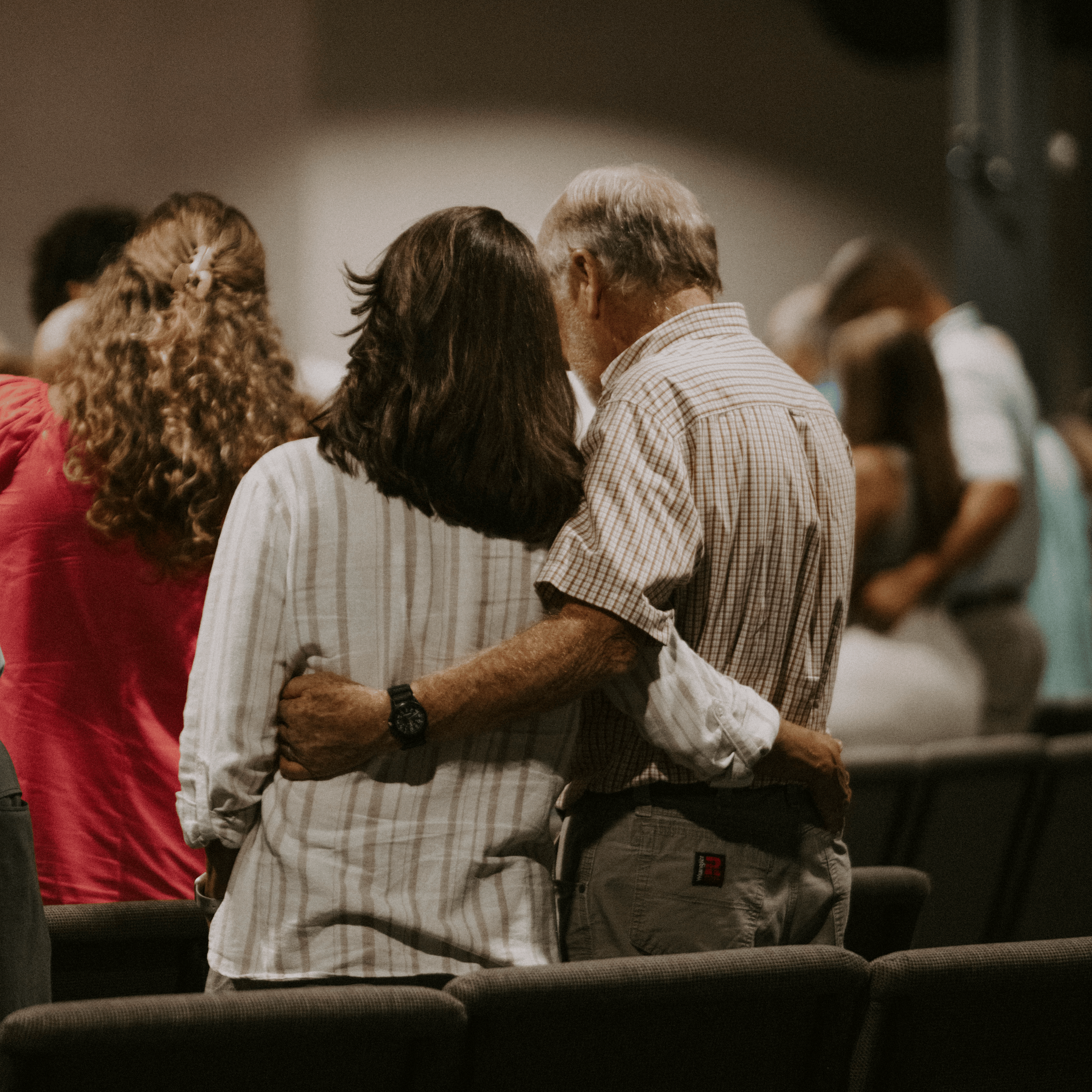 People standing closely in a circle, participating in some activity or ceremony indoors, with some holding hands and heads bowed.
