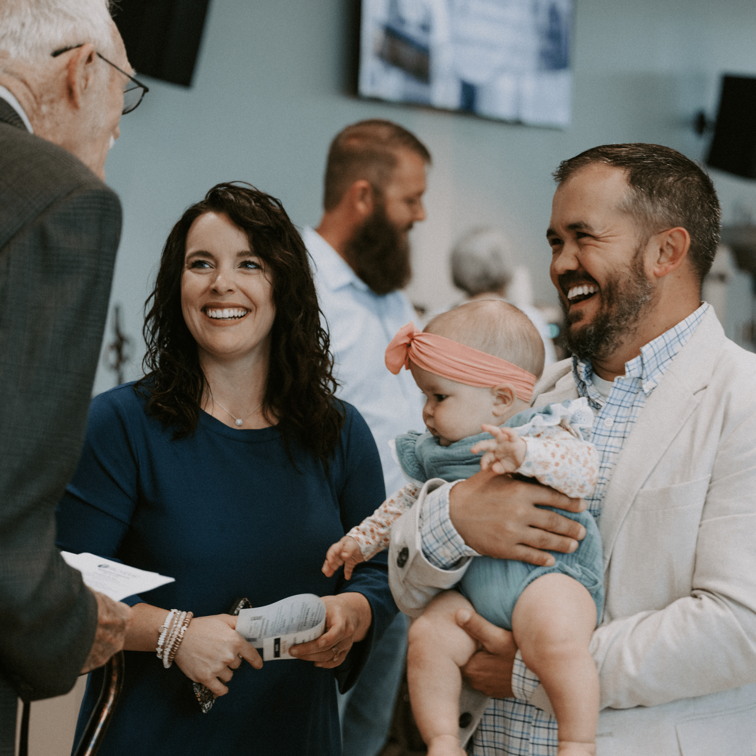A group of people at a baby shower, including a woman with long dark hair, a man with short dark hair and a beard, and an older woman with glasses, who are smiling and engaging with a baby wrapped in a blanket.