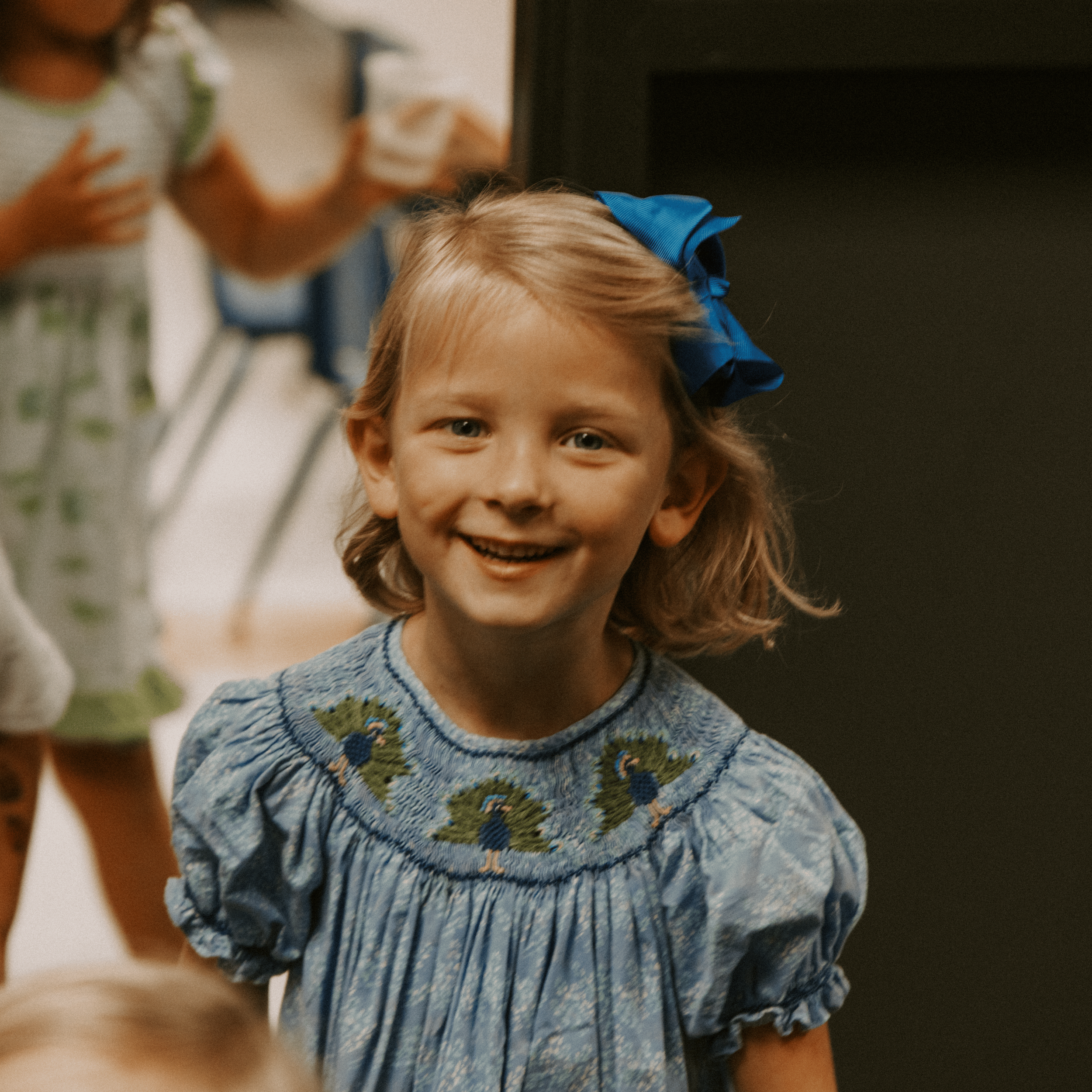 Close-up of a young girl with blonde hair and a big blue bow, smiling, wearing a blue sweater with embroidered flowers, in a room with a dark piece of furniture behind her.