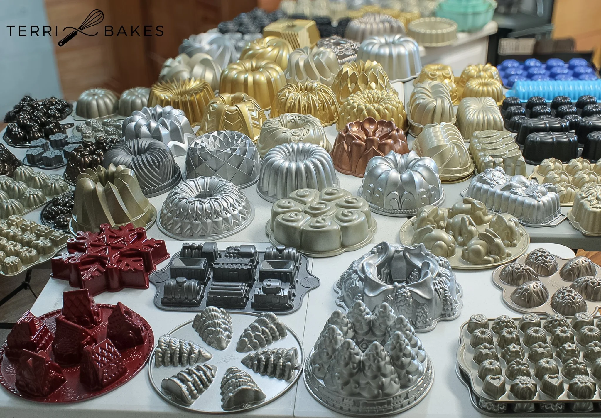 A display of various metal baking molds in different shapes and sizes, including bundt cakes, festive shapes, and decorative designs, arranged on tables at a bakery or craft store.