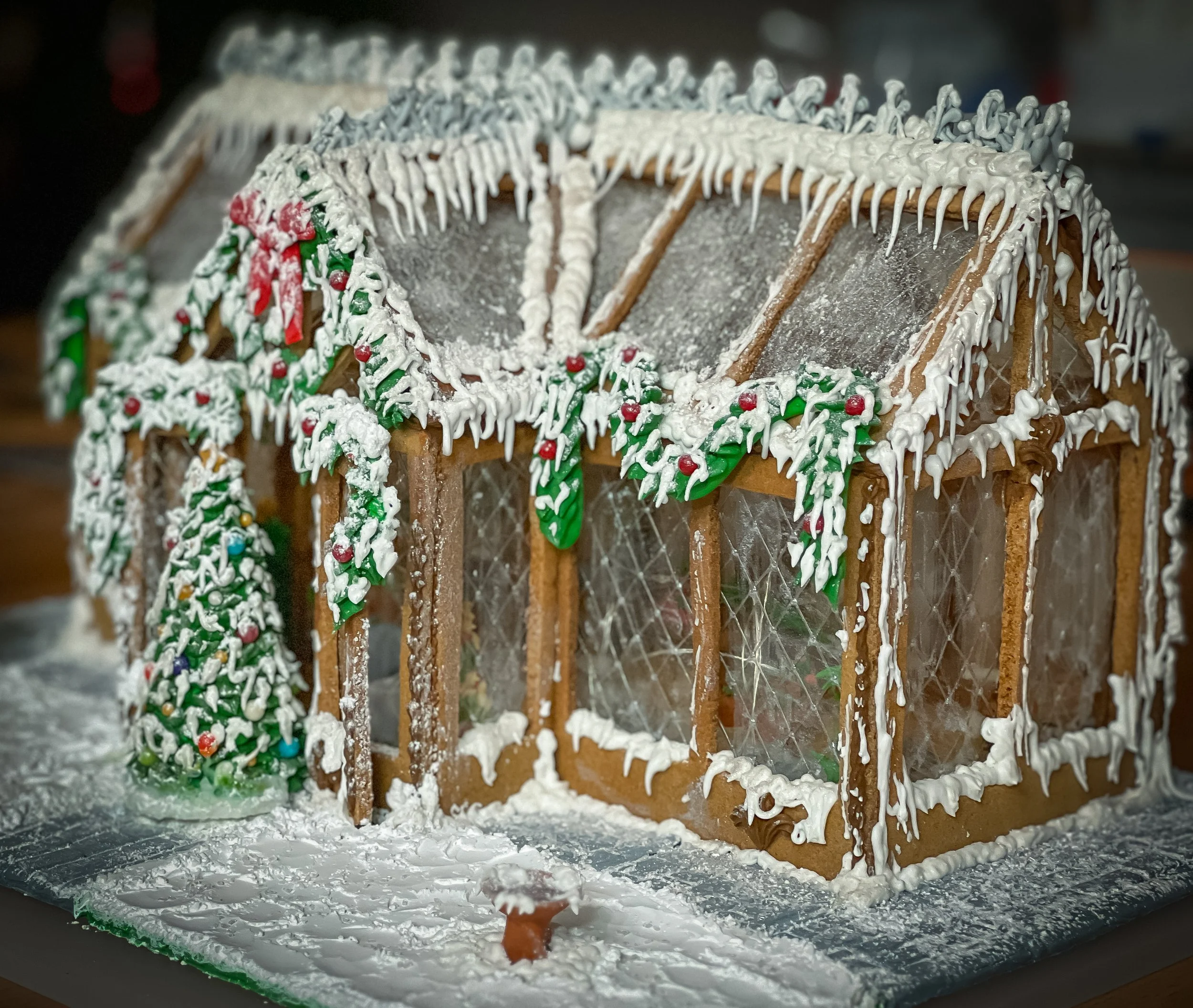 Decorative gingerbread house with white icing, green holly leaves, red berries, and a Christmas tree also decorated with icing and colorful candies.