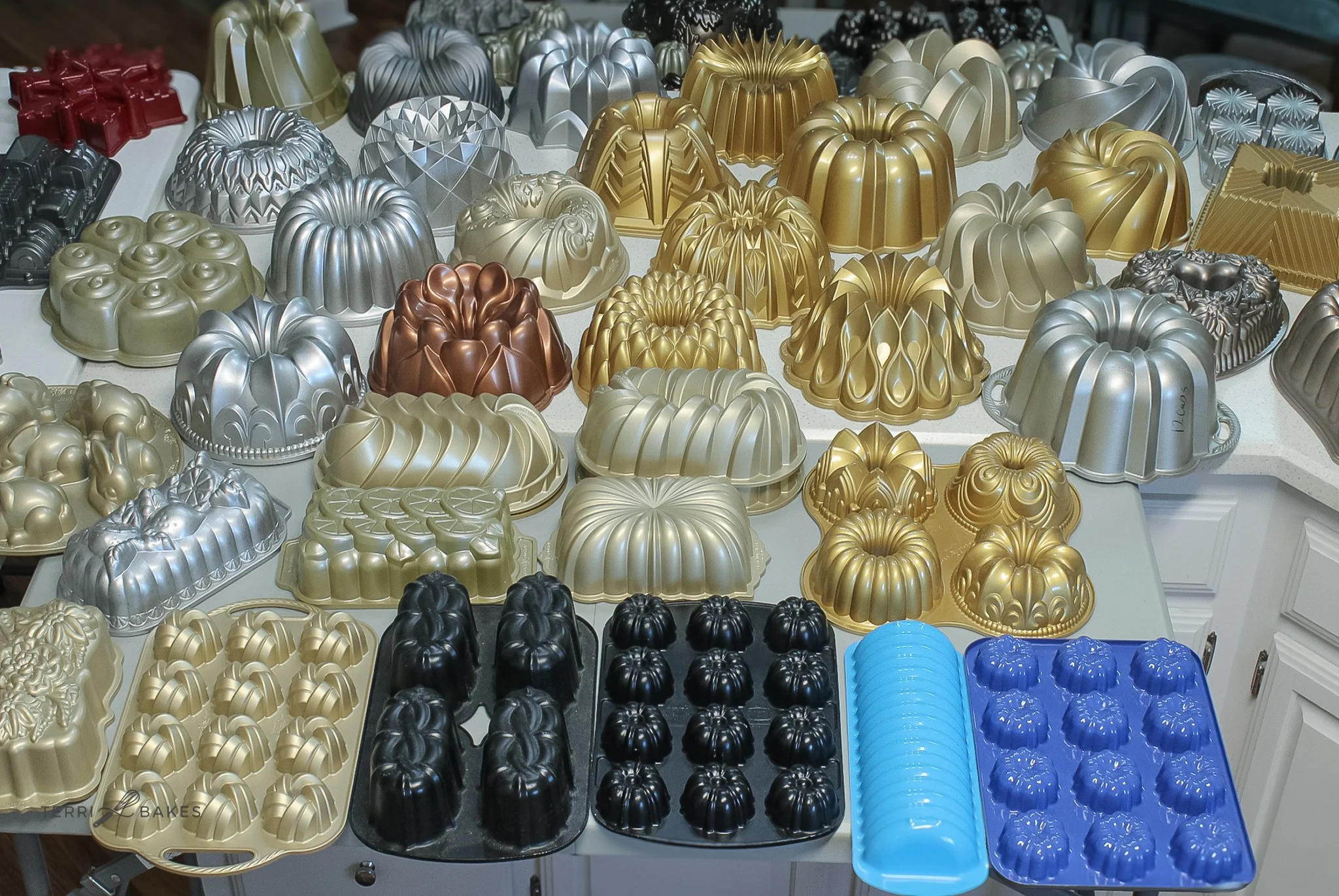 Various colorful Bundt cake pans in colors including gold, silver, black, and blue, arranged on a table.