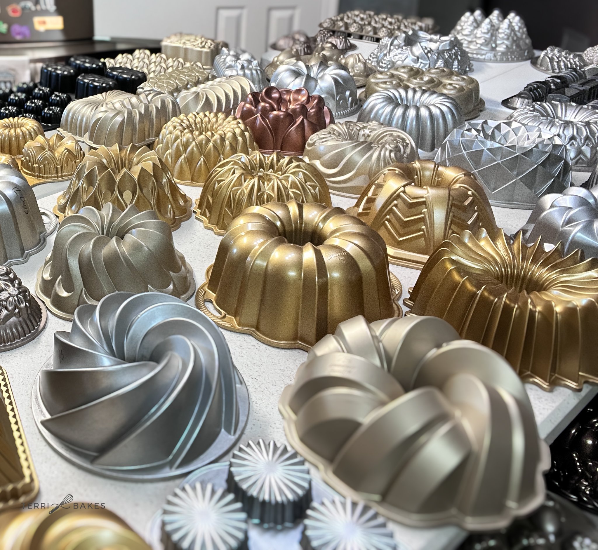 A variety of metallic Bundt pans in gold, silver, bronze, and black colors arranged on a table.