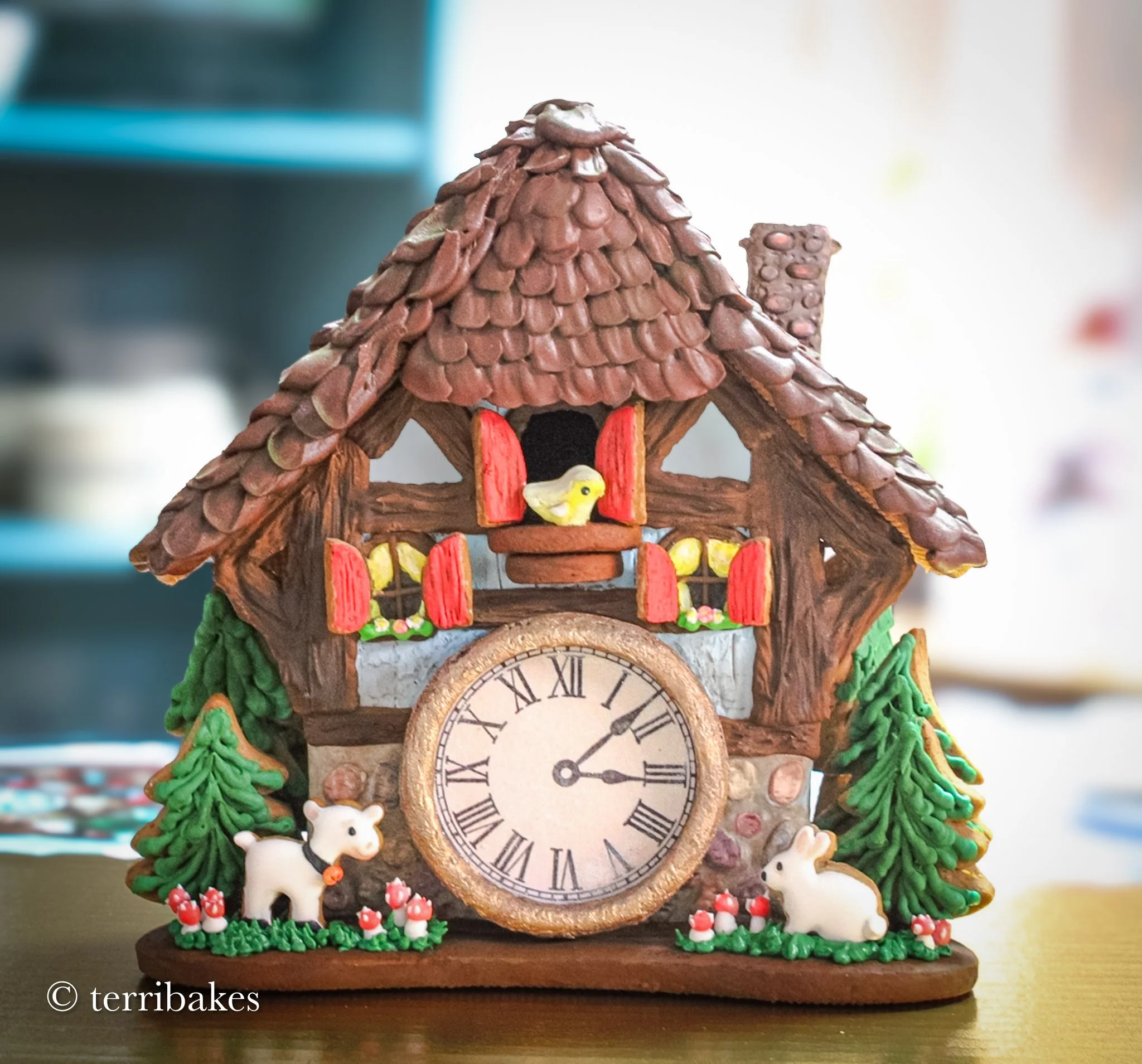 A decorative cuckoo clock featuring a brown house with a shingled roof, red window shutters, a small yellow bird in the window, and miniature trees and animals including a white rabbit and a white dog, with a clock face showing approximately 3:10