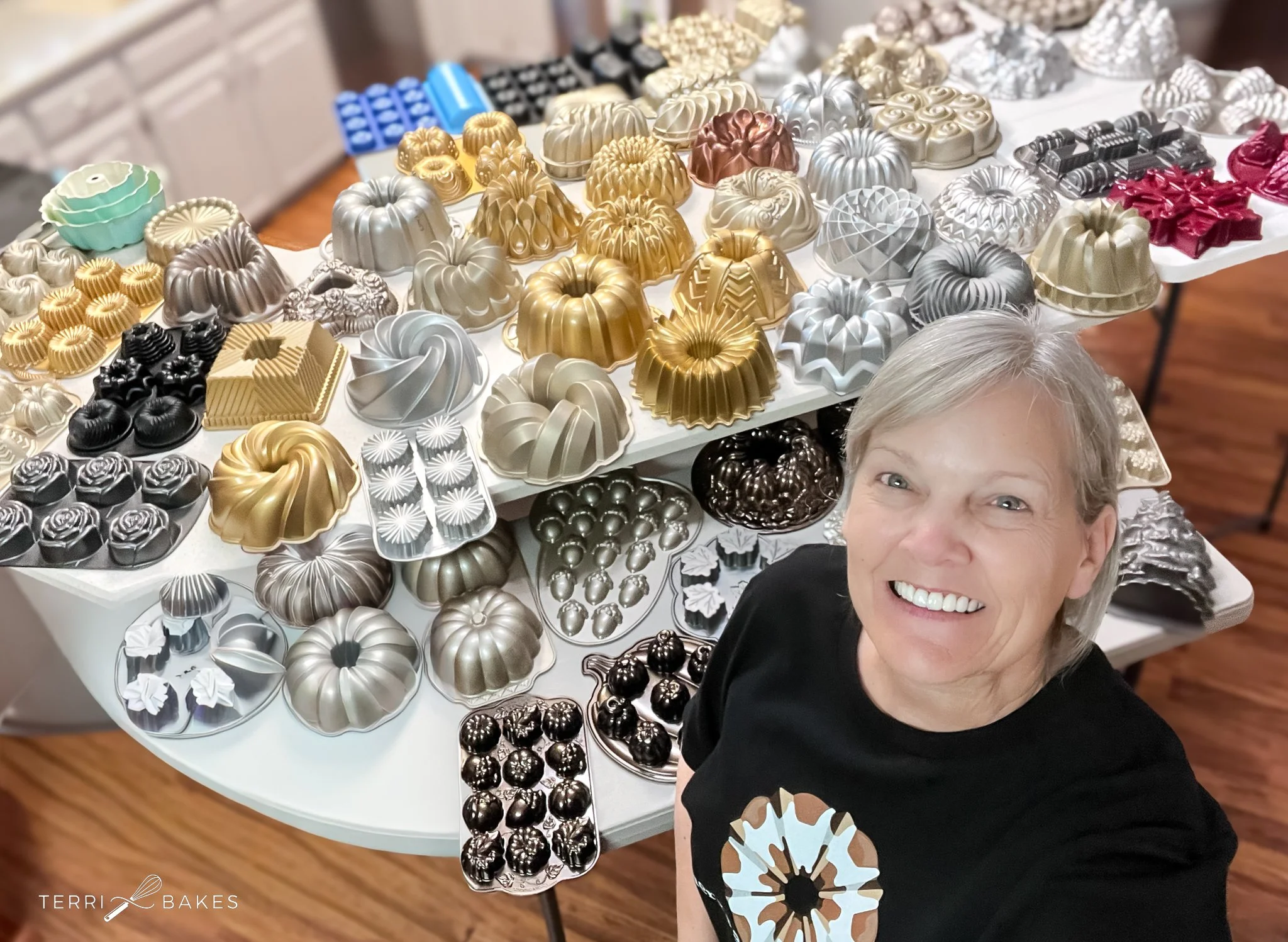A smiling woman taking a selfie in front of a table filled with a variety of colorful bundt cakes and dessert molds in metallic, black, white, gold, silver, and red colors.