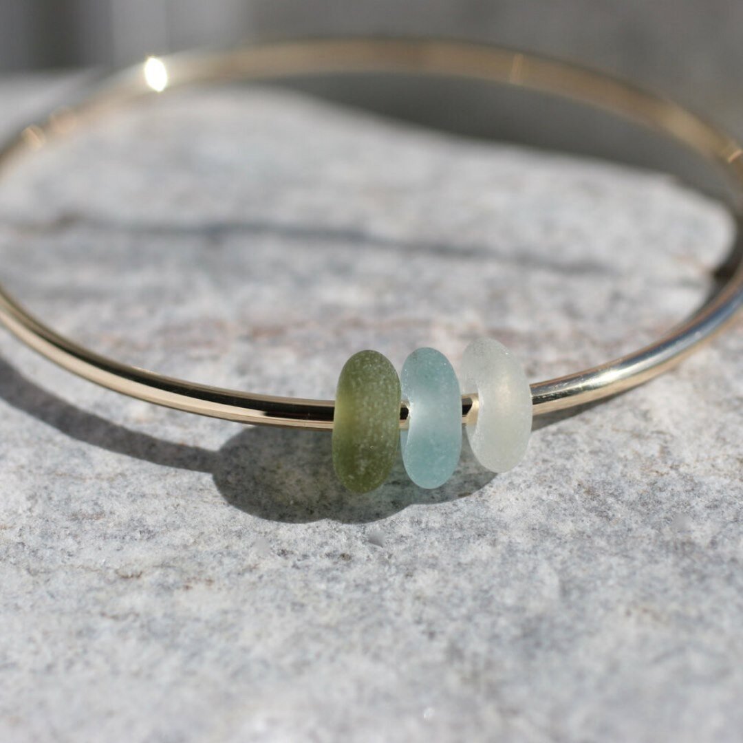 Handmade recycled 9ct yellow gold bangle strung with a trio of naturally tumbled sea glass beads found on West Country beaches. 

The bangle pictured was made in smooth recycled gold and each set of sea glass beads were individually drilled and the b