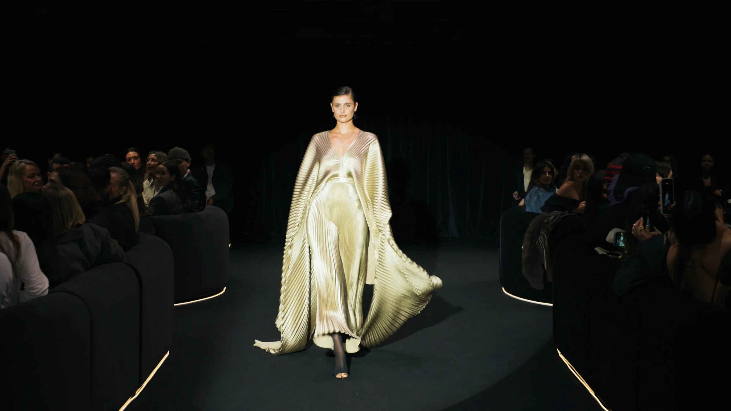 Australian Fashion Week runway cinematography featuring a gold pleated gown on a curved black runway
